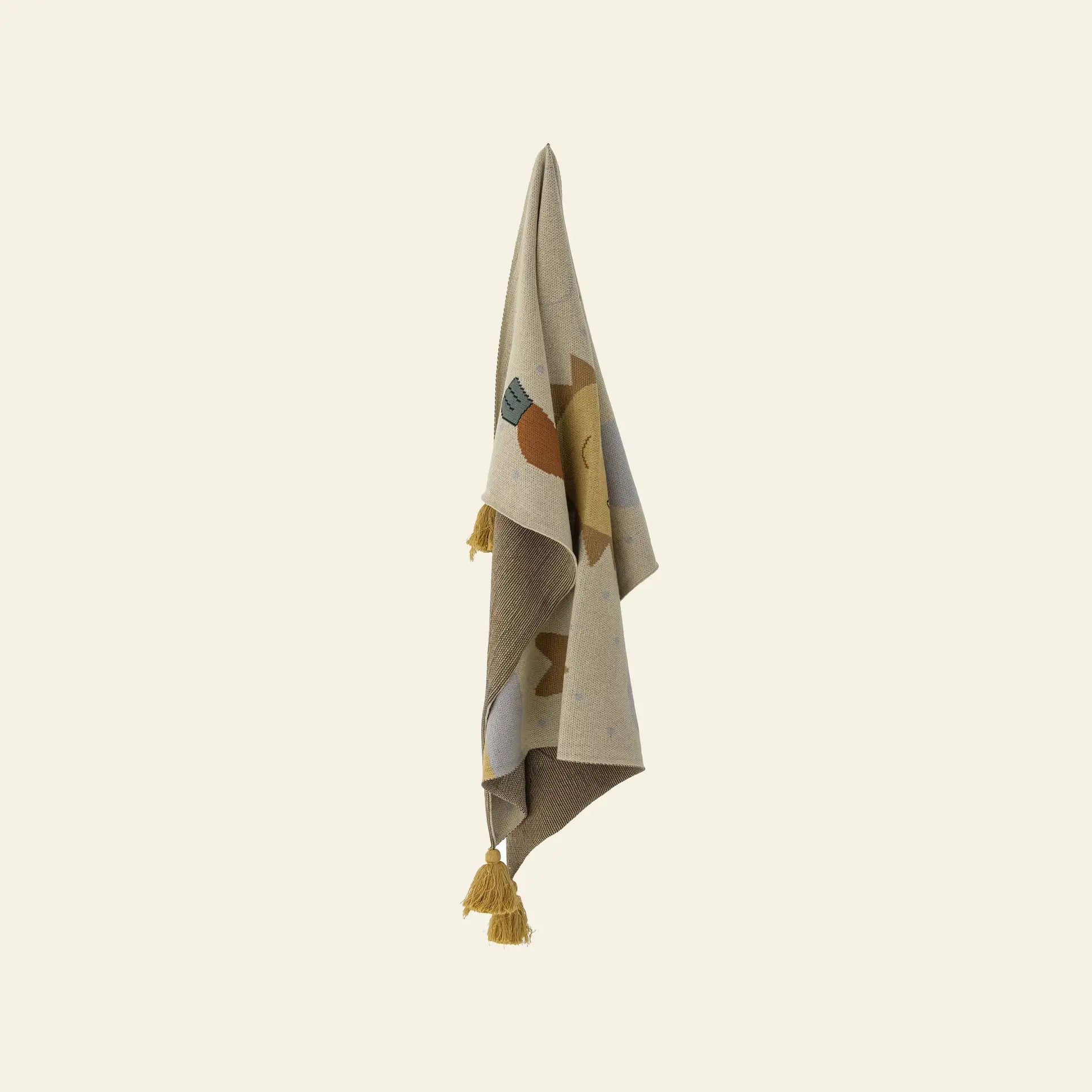 The Bloomingville Vigge Throw by Bloomingville, in beige with a colorful sun design and yellow tassels, made from OEKO-TEX certified cotton, hangs on a hook against a plain light background.