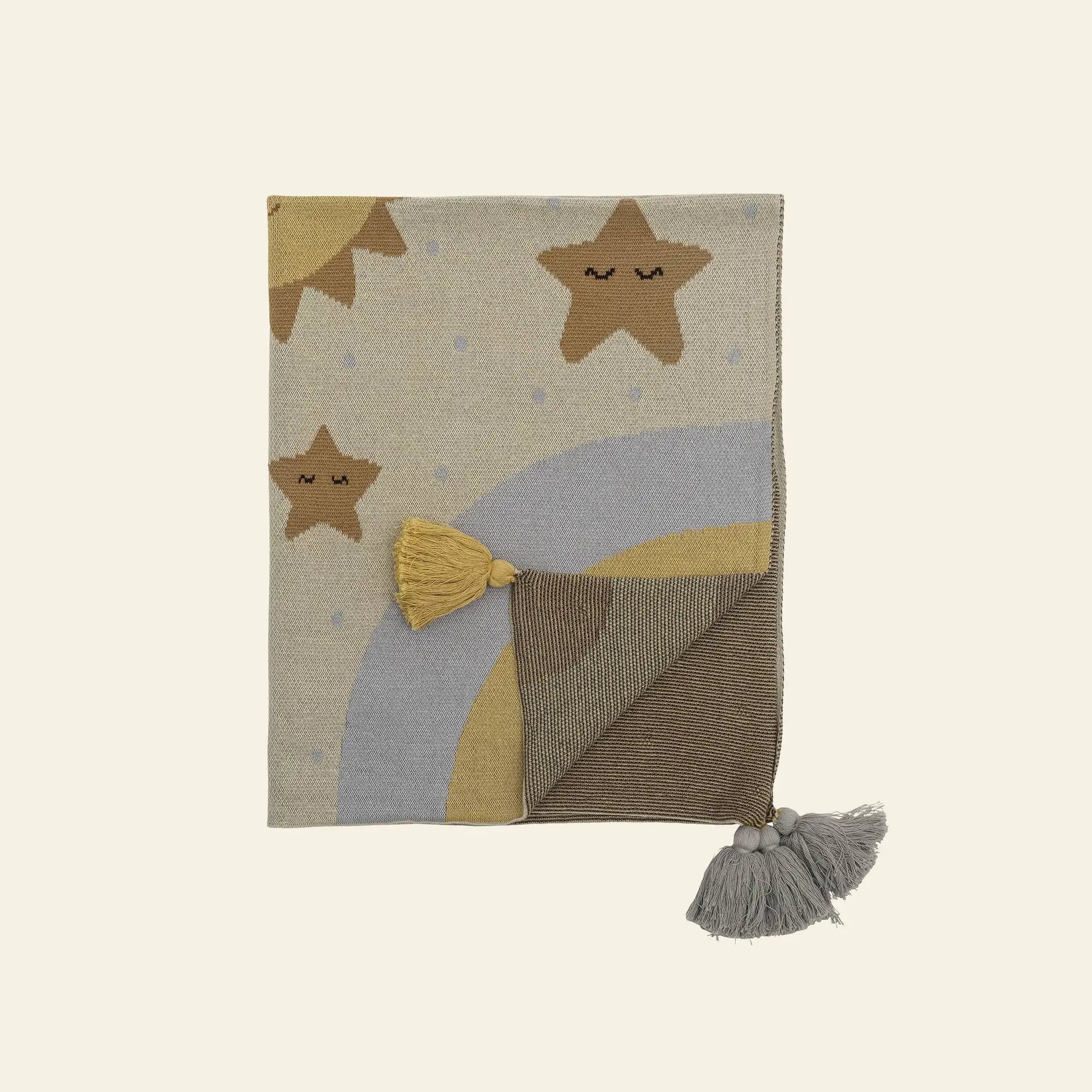 The Bloomingville Vigge Throw by Bloomingville is a folded OEKO-TEX certified cotton blanket with sky-inspired designs—smiling stars, a yellow and gray rainbow, blue dots, and corner tassels—shown on a light background.