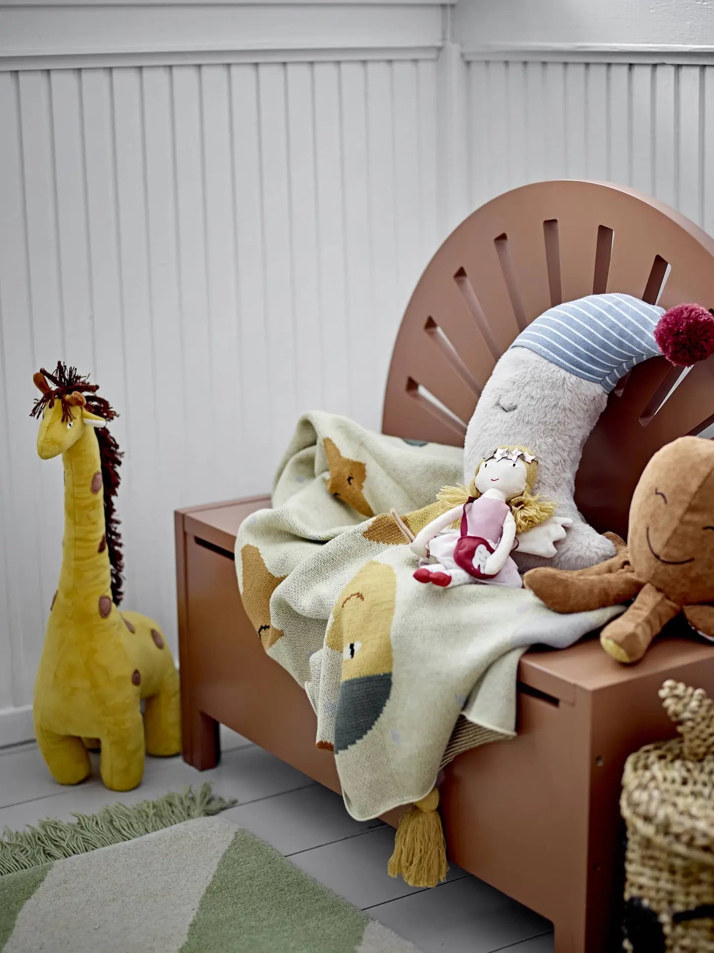 A cozy children’s room features a small brown bench with a sunburst backrest, plush toys, a doll, and the Bloomingville Vigge Throw—an OEKO-TEX certified cotton blanket with animal patterns by Bloomingville. A yellow stuffed giraffe stands nearby.