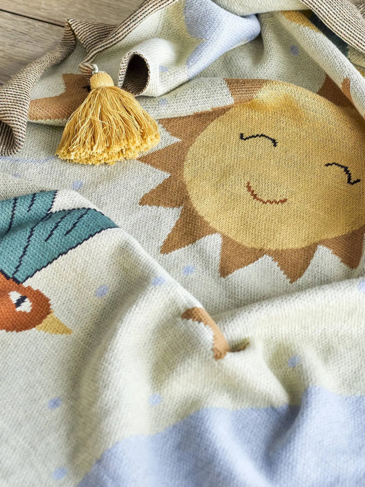The Bloomingville Vigge Throw by Bloomingville is a cozy, OEKO-TEX certified cotton knit blanket with a smiling sun, leafy patterns, blue dots, and a yellow tassel in one corner for added charm and a sky-inspired touch.