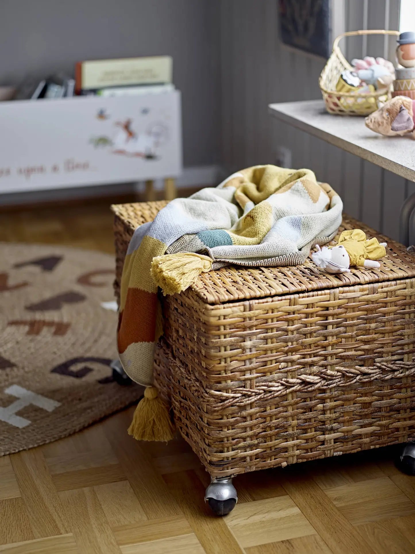 A wicker storage box with wheels sits on a wooden floor. The Bloomingville Vigge Throw, a striped OEKO-TEX certified cotton blanket by Bloomingville, is draped over it, with a small stuffed toy on top amid cozy neutral décor and kids furniture.