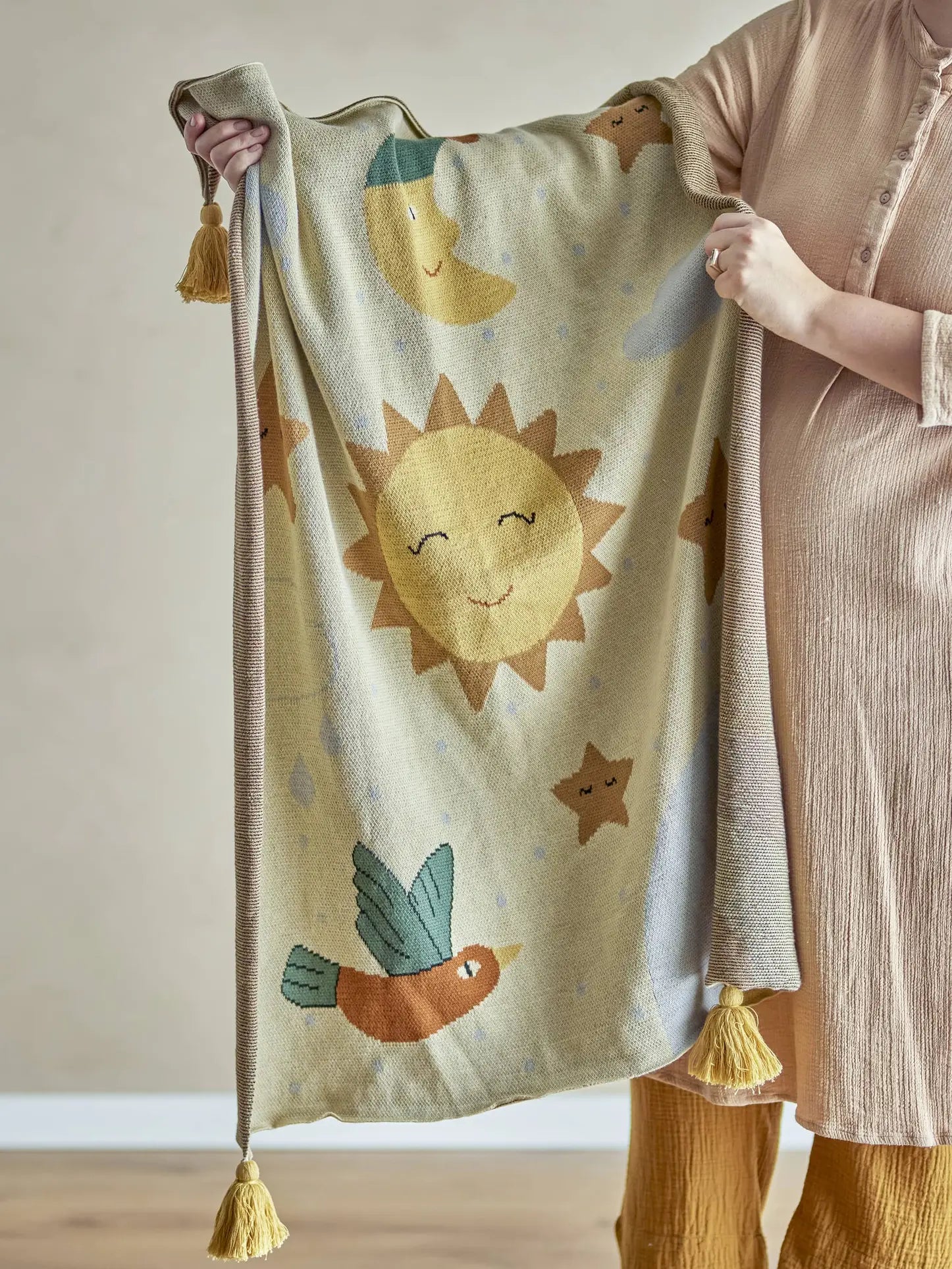 A person in neutral, loose clothing holds up the Bloomingville Vigge Throw—a soft yellow knitted blanket from Bloomingville, crafted with OEKO-TEX certified cotton and decorated with sky-themed designs and yellow tassels on the corners.