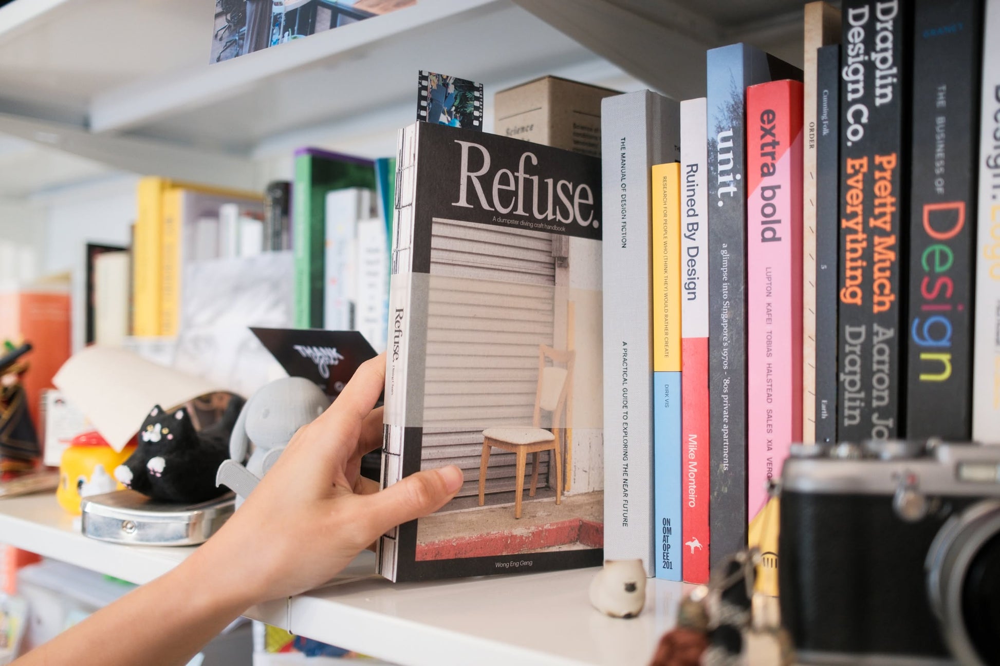 A hand pulls Bluetaped Refuse: A Dumpster Diving Craft Handbook by Wong Eng Geng from a shelf lined with colorful books, a black cat figurine, and a camera—capturing Bluetaped’s creative upcycling spirit.