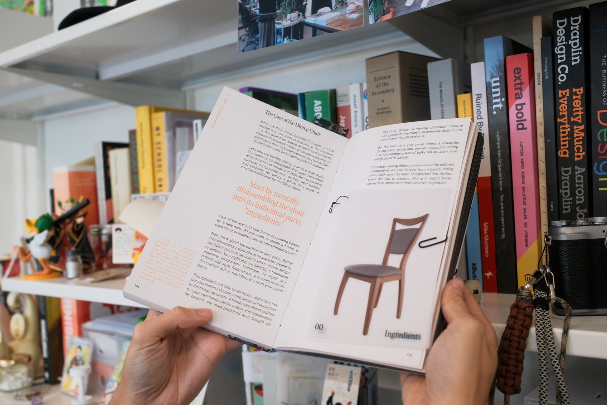 Someone holds open Bluetaped Refuse: A Dumpster Diving Craft Handbook by Wong Eng Geng (Bluetaped), showing an Ingredients section and a photo of an upcycled wooden chair, in front of a bookshelf filled with colorful books and objects.