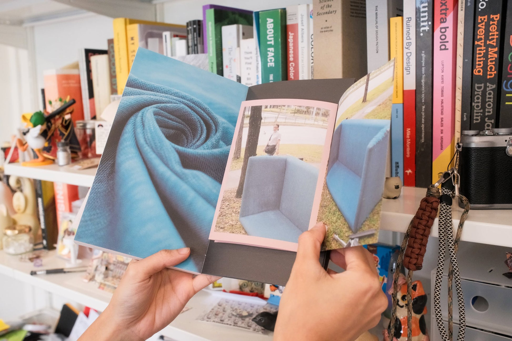 Two hands hold open Bluetaped Refuse: A Dumpster Diving Craft Handbook by Wong Eng Geng (Bluetaped), displaying photos of upcycled blue fabric and a blue, cube-shaped chair. In the background is a white bookshelf with books and assorted items.