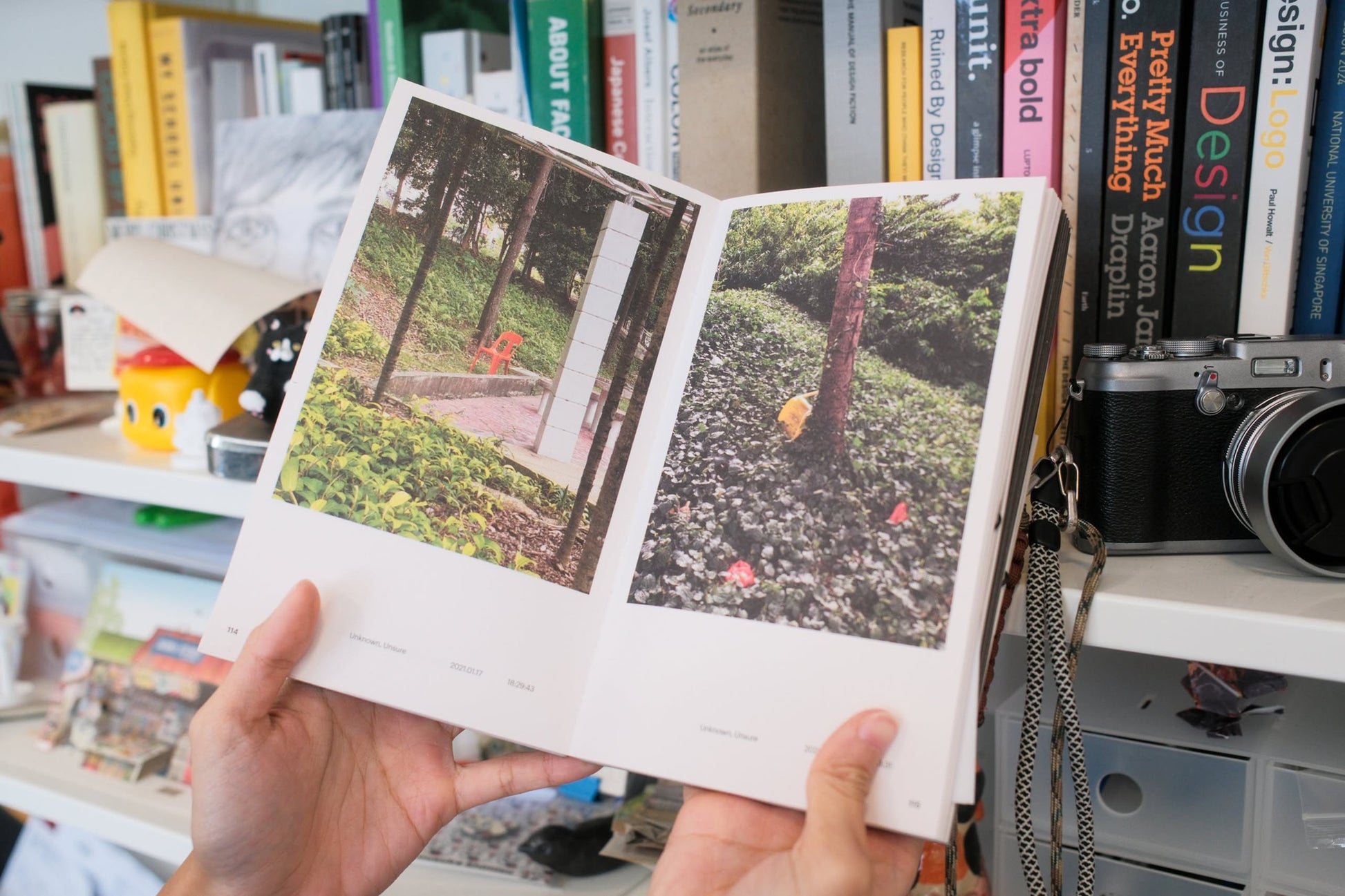 A person holds open Bluetaped Refuse: A Dumpster Diving Craft Handbook by Wong Eng Geng from Bluetaped, displaying outdoor photos amid trees in front of a cluttered bookshelf filled with books, a camera, and upcycled furniture waste.