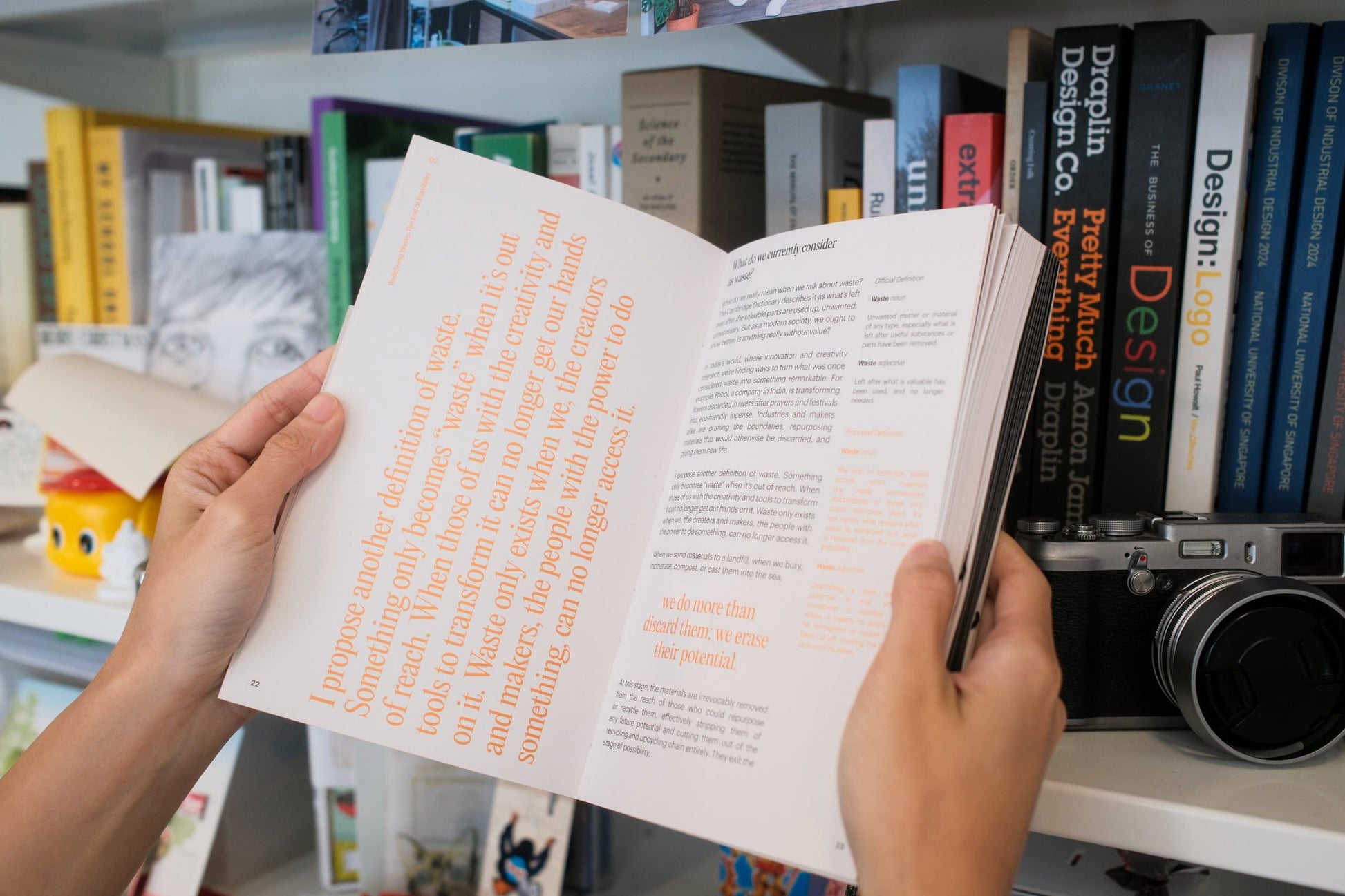 A person holds open Bluetaped Refuse: A Dumpster Diving Craft Handbook by Wong Eng Geng (Bluetaped), standing before a bookshelf filled with books, a camera, and upcycled objects made from furniture waste.