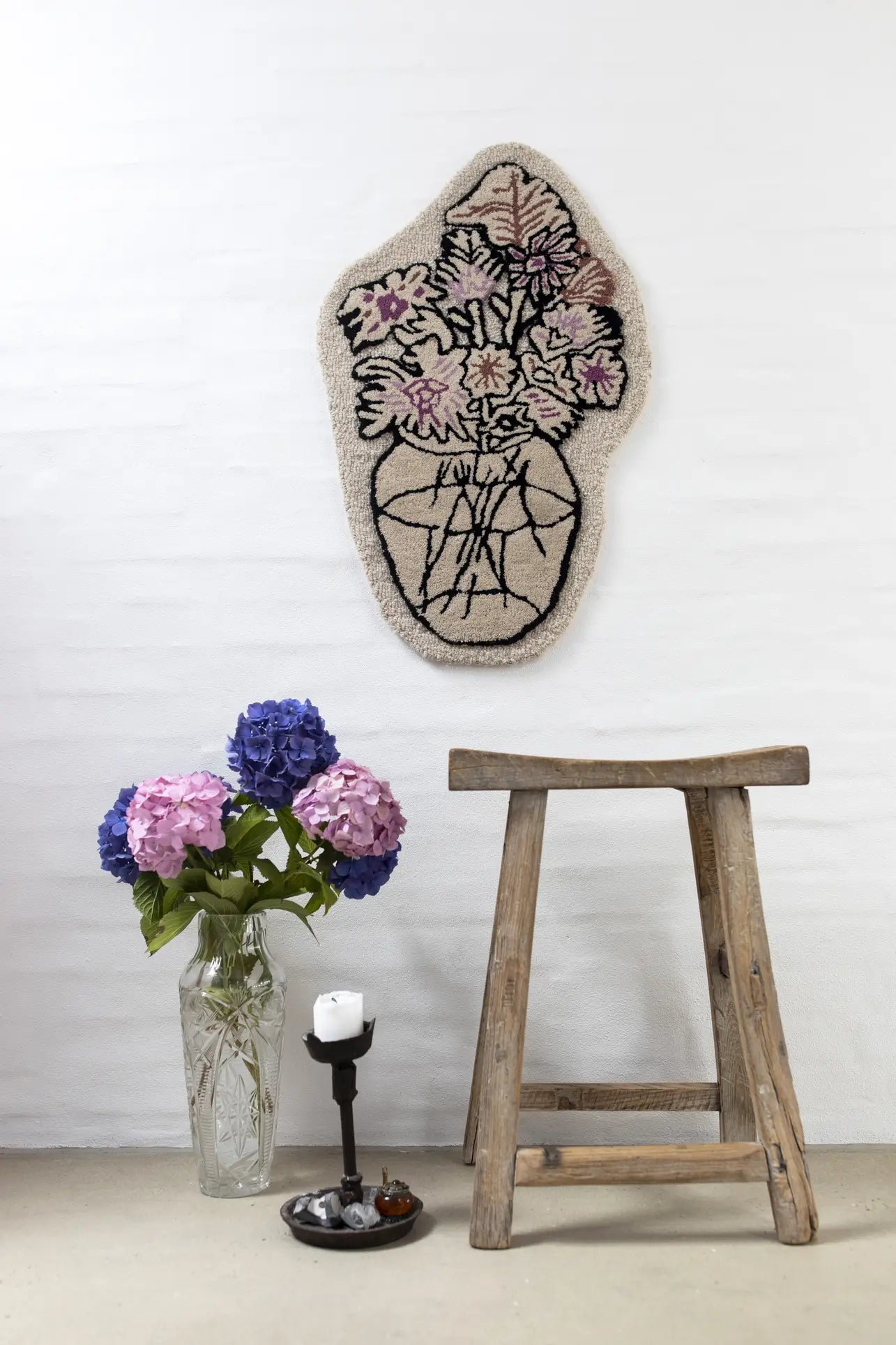 A rustic wooden stool sits by a glass vase of purple and pink hydrangeas; above, the Bongusta Autumn Flowers Rug by Bongusta, crafted from 100% wool, displays a floral vase design against a white brick wall.