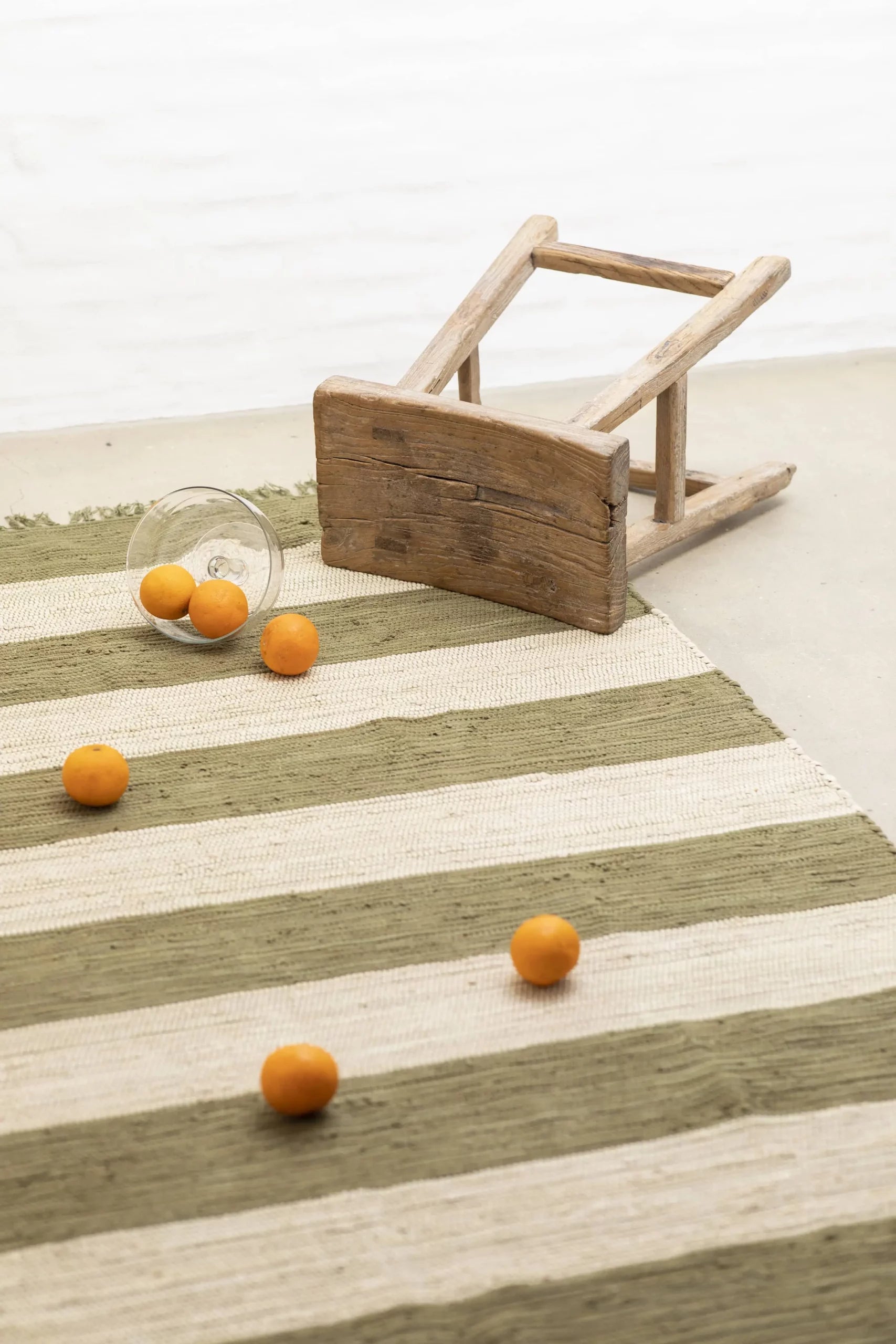 A small wooden stool tipped over on a Bongusta Chindi Rug (45x60) by Bongusta, with several orange fruits and a glass cup spilled on the floor.