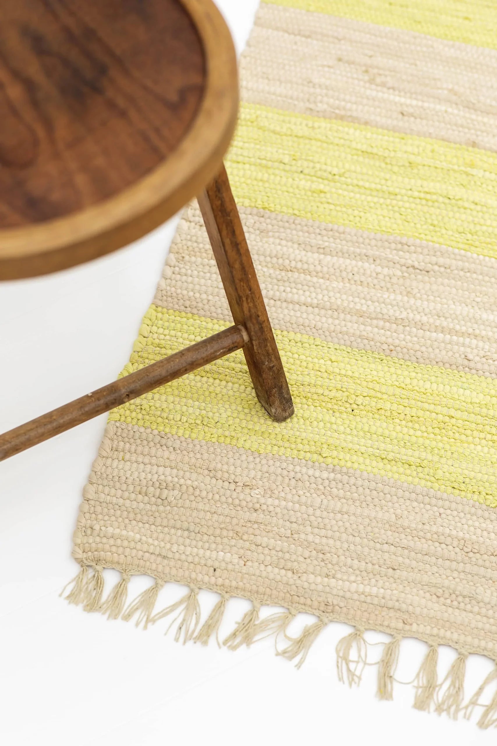 A wooden stool stands on the Bongusta Chindi Rug (45x60) by Bongusta, handcrafted in India from surplus cotton with beige and yellow stripes and fringed edges. The photo, shot from above, shows part of the stool and a corner of the rug.