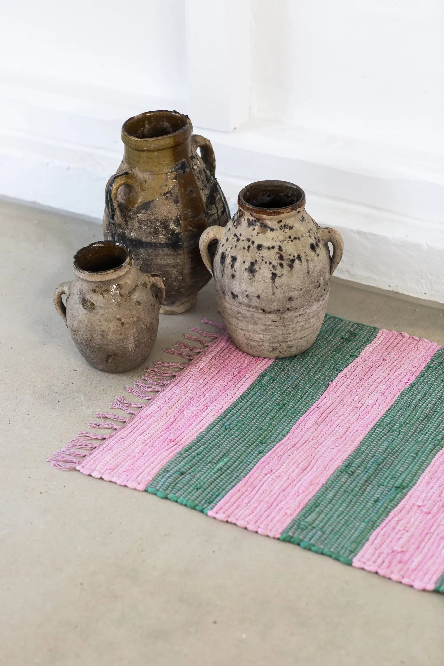 Three rustic earthenware jars with handles sit on a light floor beside the Bongusta Chindi Rug (45x60) by Bongusta, handcrafted in India from surplus cotton with pink and green stripes and fringe. The backdrop is a white wall.