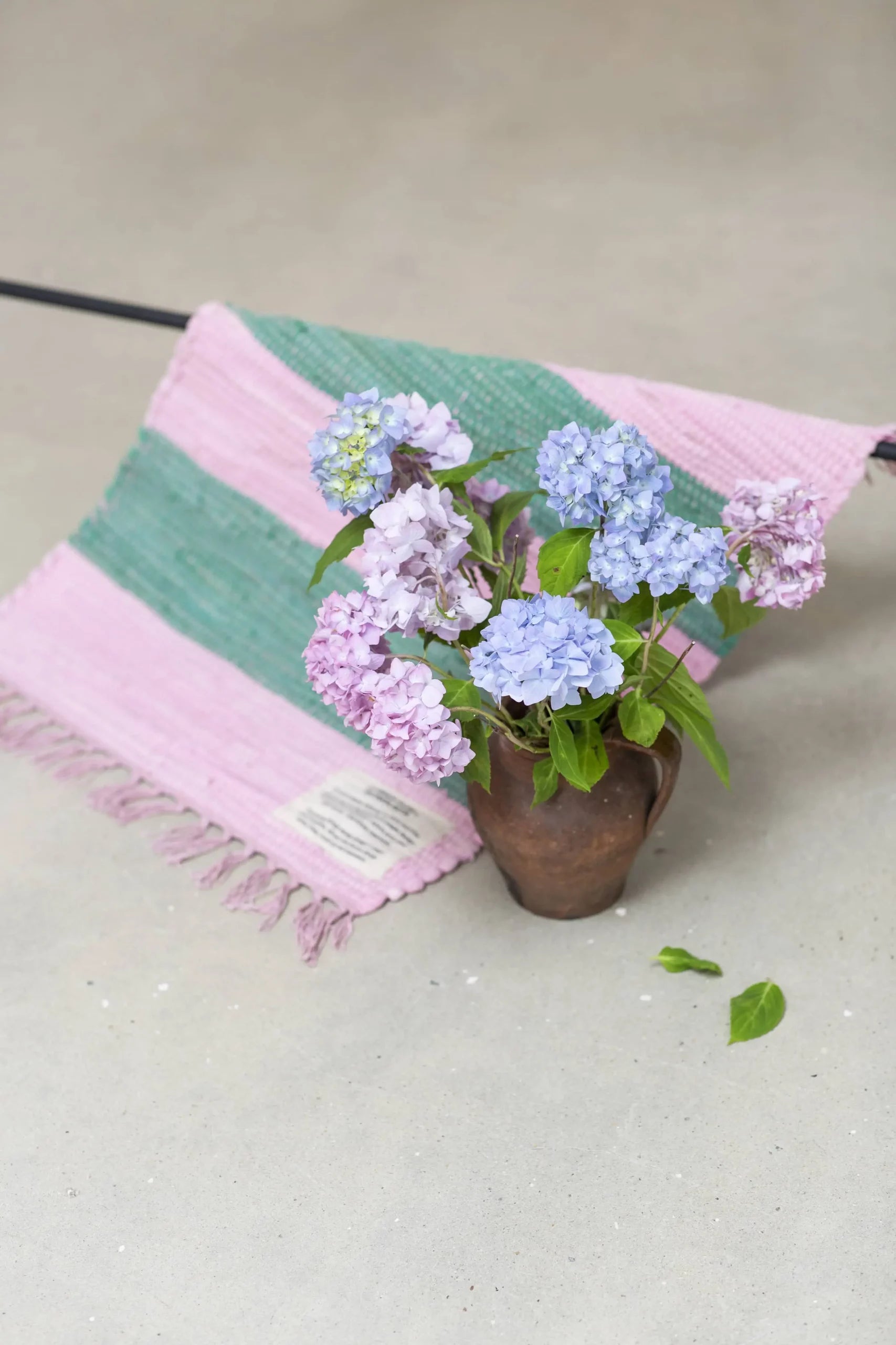 A vase with light purple and blue hydrangeas sits on a light gray surface beside the Bongusta Chindi Rug (45x60) by Bongusta, handcrafted in India from surplus cotton, featuring pink and green stripes, fringed edges, and a small label.