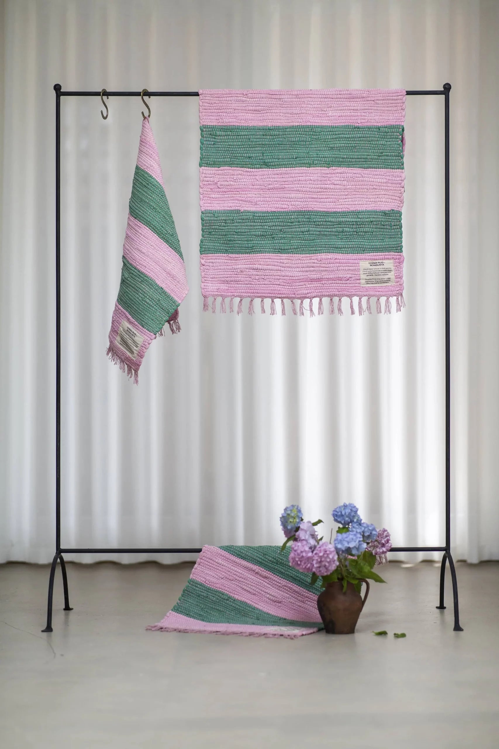 A black metal rack displays two Bongusta Chindi Rugs (45x60) by Bongusta, handcrafted in India from surplus cotton with pink and green stripes; a matching rug is beside a brown vase of hydrangeas. White curtains hang behind.