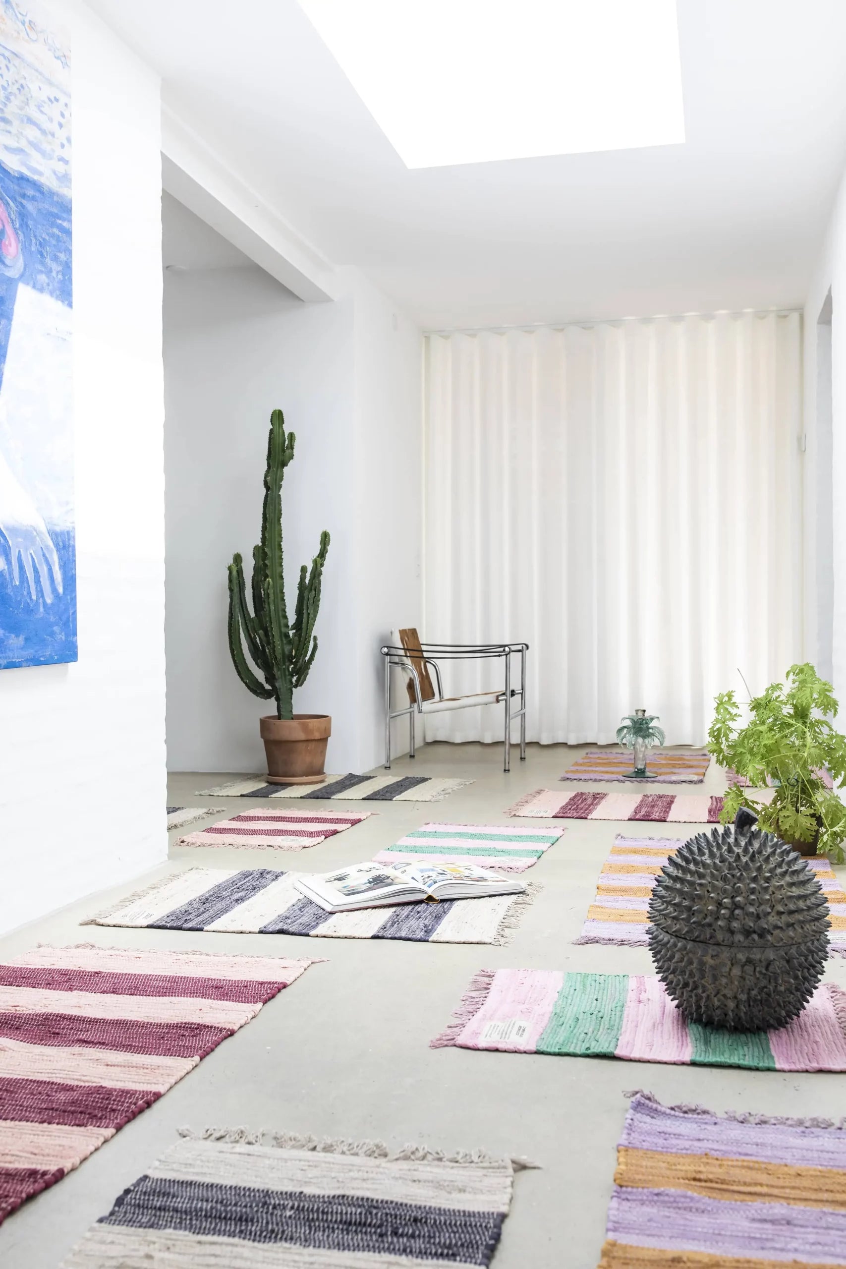 A bright hallway lit by a skylight features the Bongusta Chindi Rug (45x60) by Bongusta, handcrafted in India from surplus cotton, plus a tall potted cactus, green plants, a spiky black sculpture, white curtains, and a blue painting on the wall.
