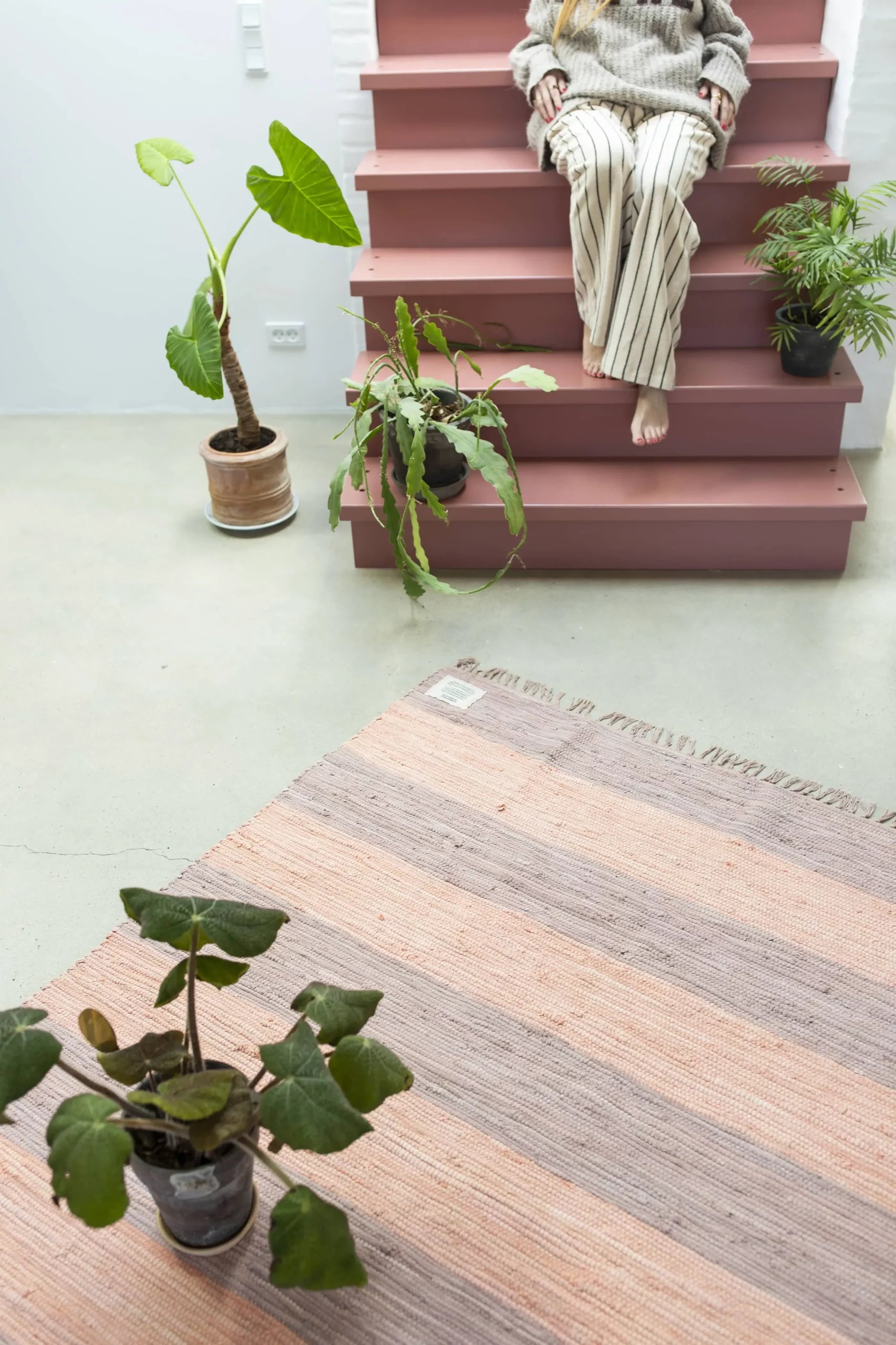A person in striped pants sits barefoot on pink stairs with potted plants and the Bongusta Chindi Rug (45x60) by Bongusta, handcrafted in India from surplus cotton, adding color to a bright minimalist room with more greenery on the floor.