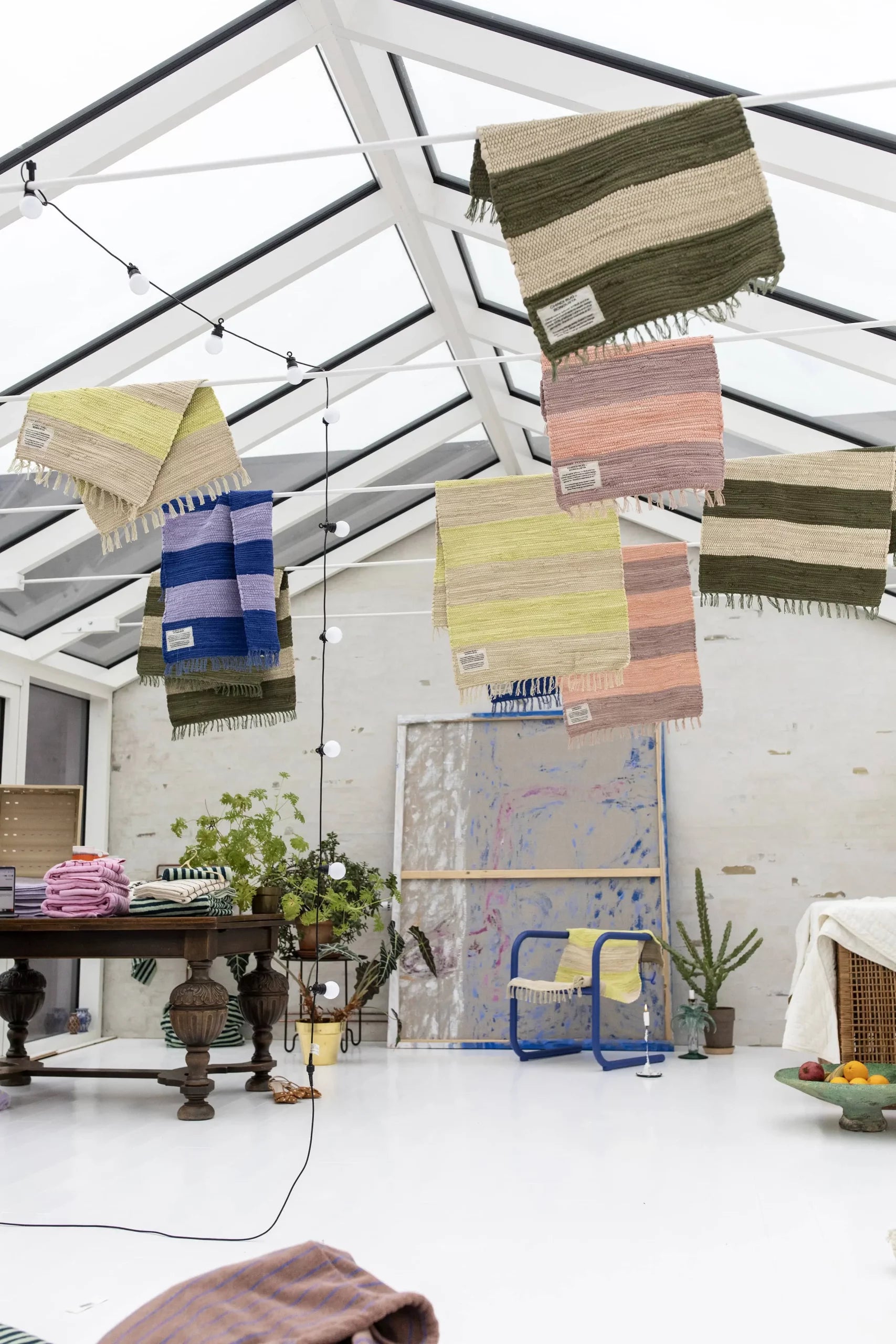 A Bongusta Chindi Rug (45x60), handcrafted in India, hangs in a bright sunroom with colorful woven blankets. Potted plants, a rustic wooden table, abstract art, and cotton fabric accents complete the eclectic, airy decor.