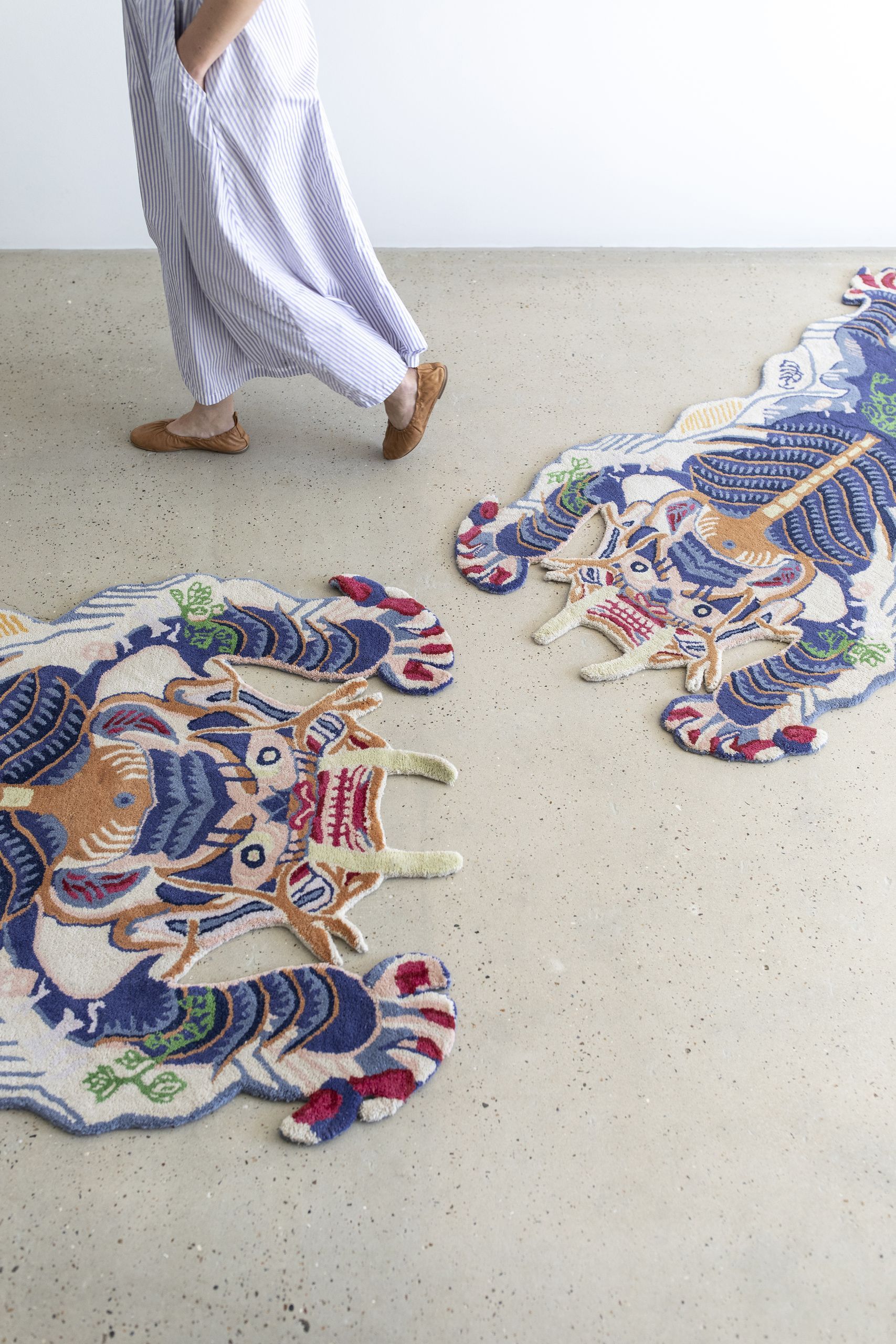 A person in a striped dress and brown shoes walks past two small Bongusta Dragon Rugs by Bongusta, featuring colorful, intricate patterns on a light, speckled concrete floor.