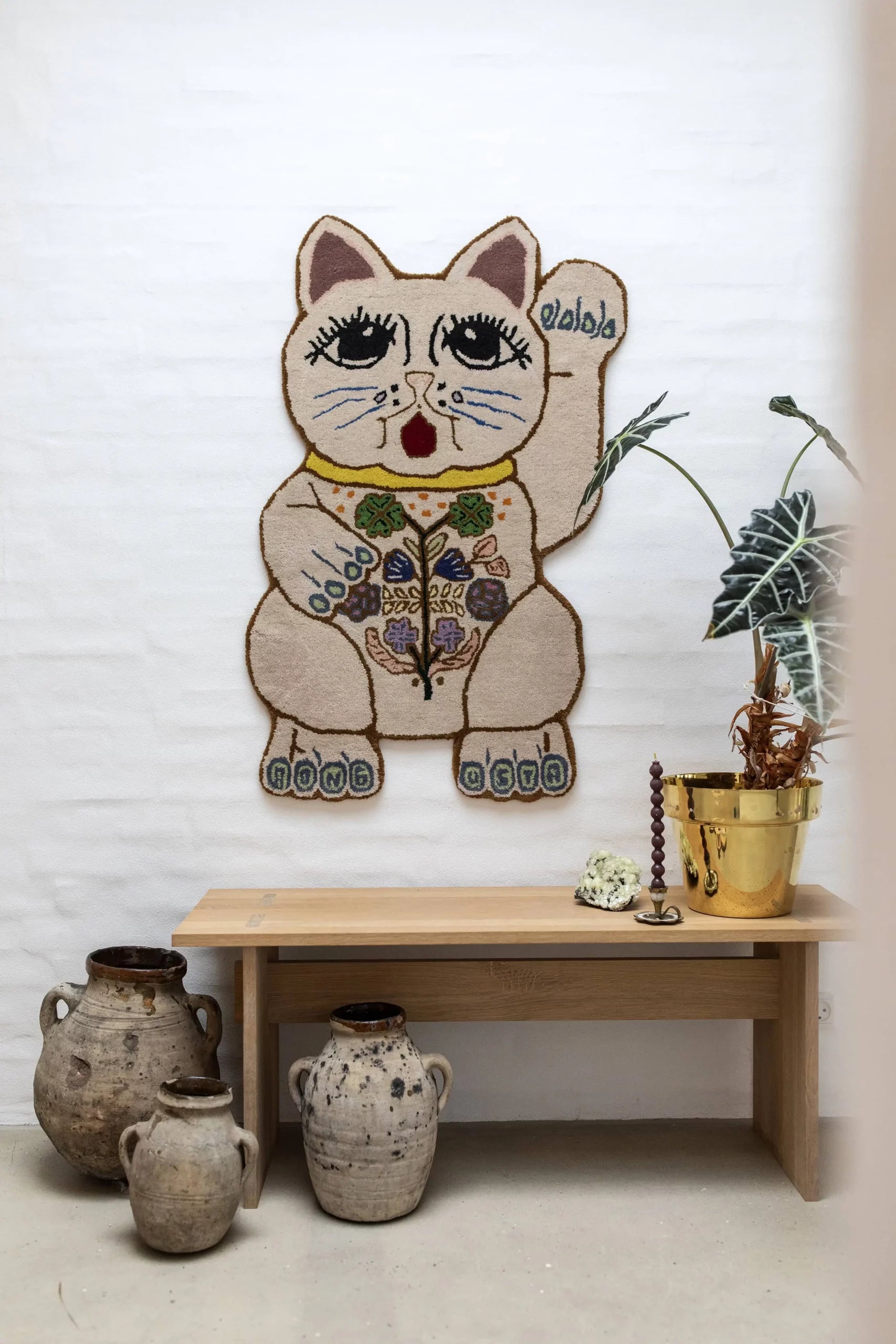 The Bongusta Lucky Rug by Bongusta, featuring big eyes and a raised paw, hangs above a wooden bench. The bench displays a gold pot with greenery and a small sculpture, with rustic ceramic jugs beside it. Subtle floral accents complete the look.