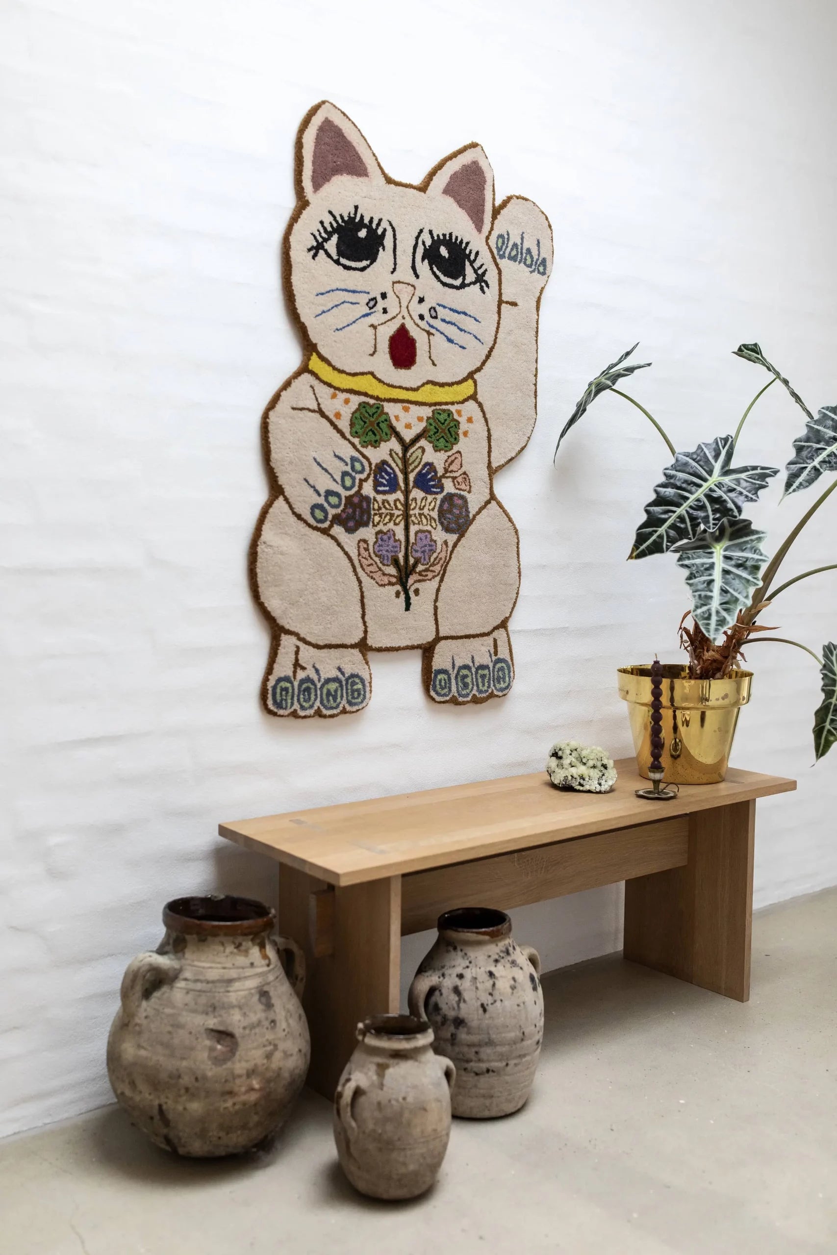 The Bongusta Lucky Rug by Bongusta hangs on a white wall above a light wooden bench, paired with a gold planter holding a leafy plant and three rustic clay pots placed on the floor.