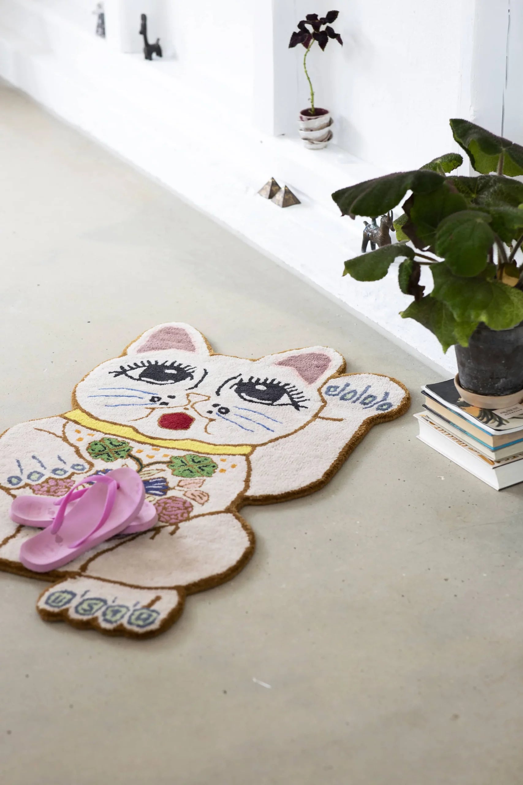 The Bongusta Lucky Rug, hand-tufted from wool, features floral accents on its cat-shaped design. Pink flip-flops rest on it while a potted plant atop stacked books sits nearby against a white wall.