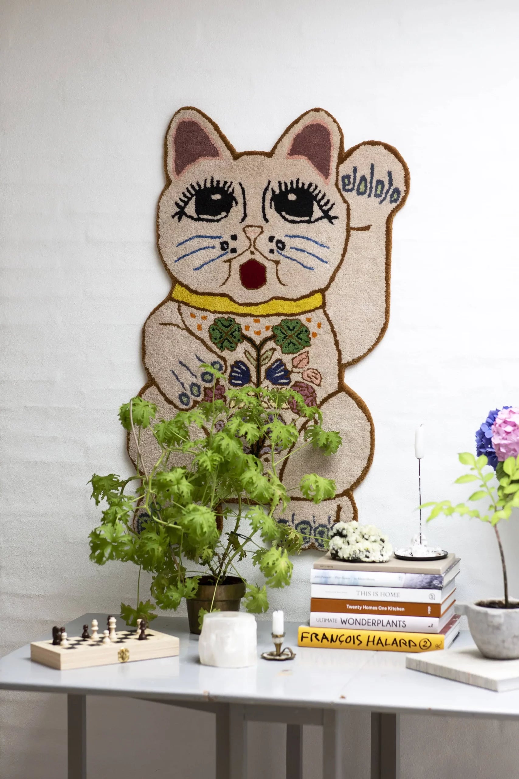 The Bongusta Lucky Rug, hand-tufted in wool with floral details, hangs above a table decorated with potted plants, books, candles, a chess set, and purple flowers against a white brick wall.