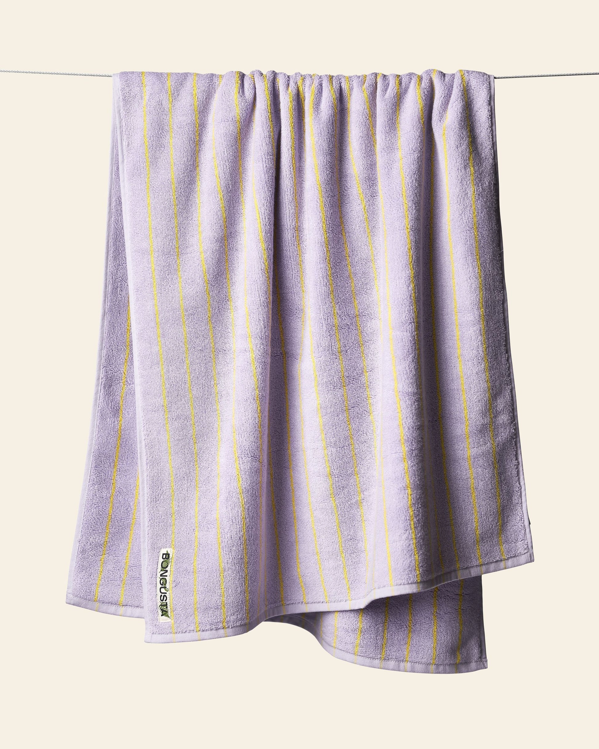 A Bongusta Naram Bath Towel in lavender with thin yellow stripes hangs on a clothesline against a light beige background. Made from Oeko-Tex combed cotton, this luxury towel by Bongusta displays a small white tag in one corner.