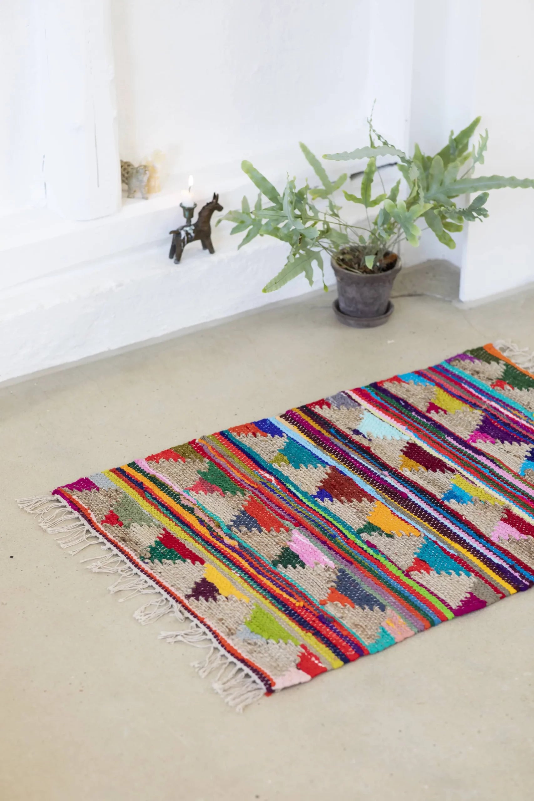 The Bongusta Pyramid Chindi Rug (60x90), woven with cotton and jute, brightens the room beside a green potted plant and white wall, adding a cozy touch to the space.