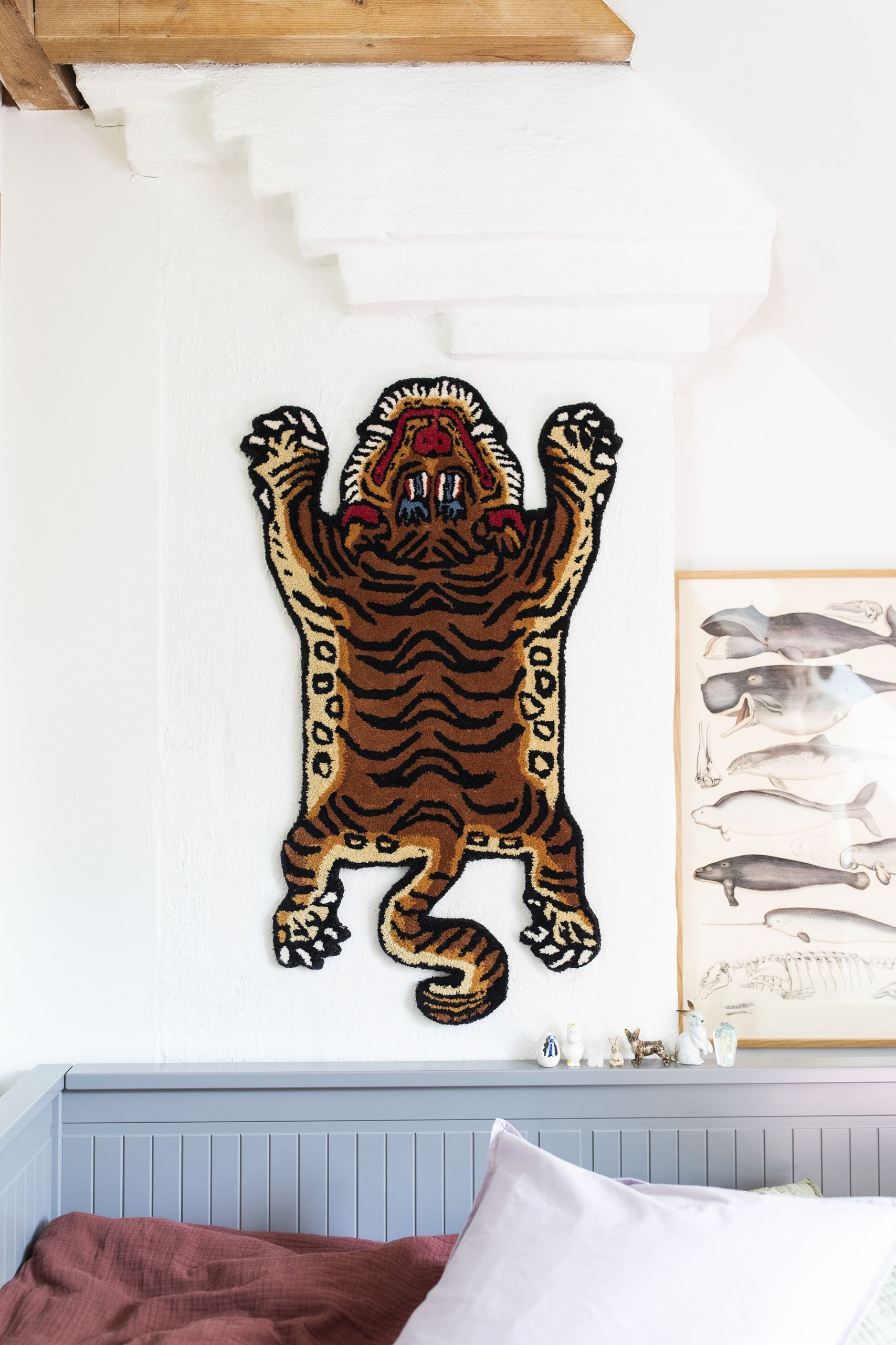A Bongusta Tiger Rug, Baby by Bongusta is mounted on a white wall under a staircase next to a framed animal illustration, creating a jungle vibe. Below, part of a gray bed with a red blanket can be seen.