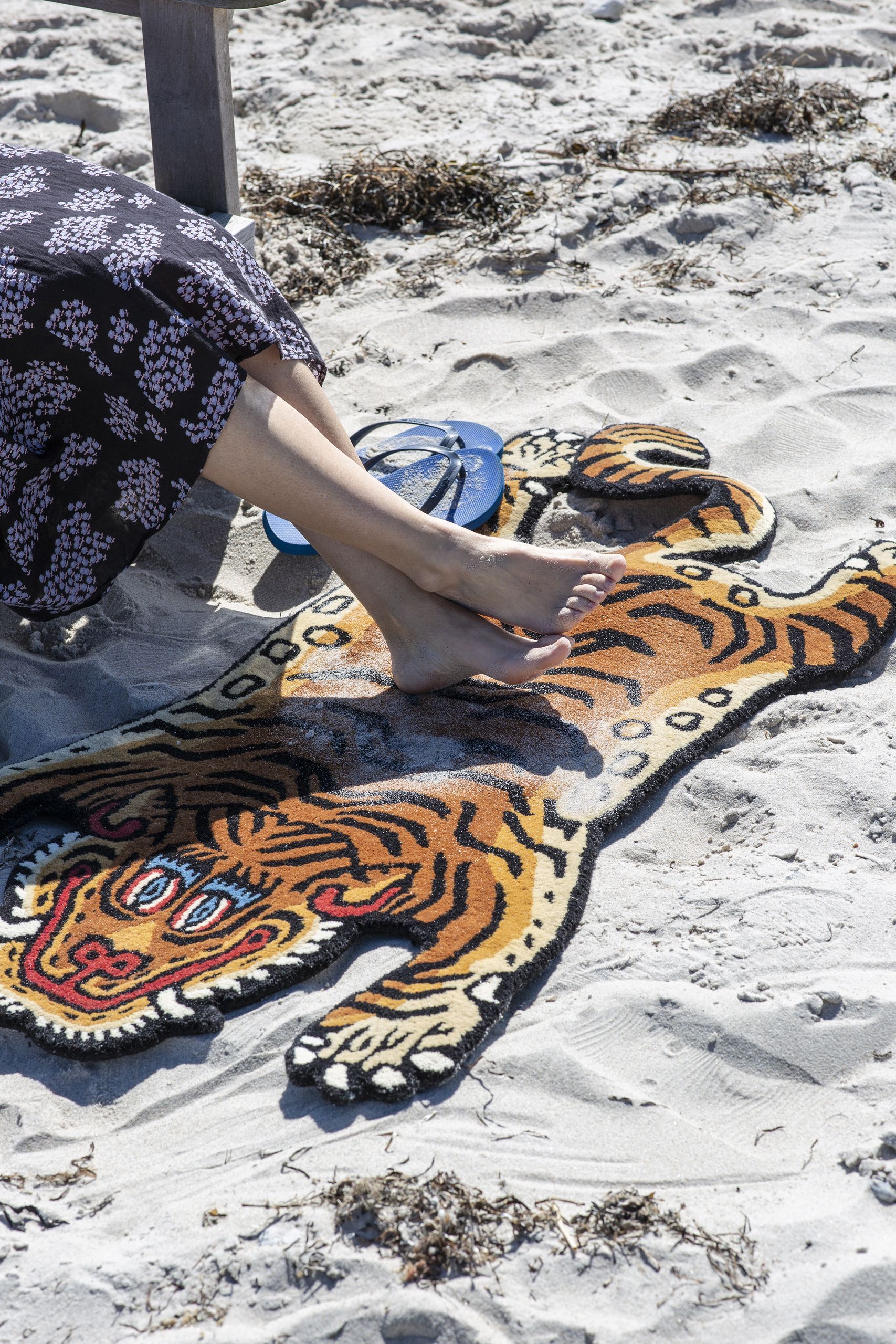 A person in a patterned skirt sits barefoot on the sand, resting their feet on the Bongusta Tiger Rug, Baby by Bongusta. Blue sandals lie nearby, and seaweed is scattered around.