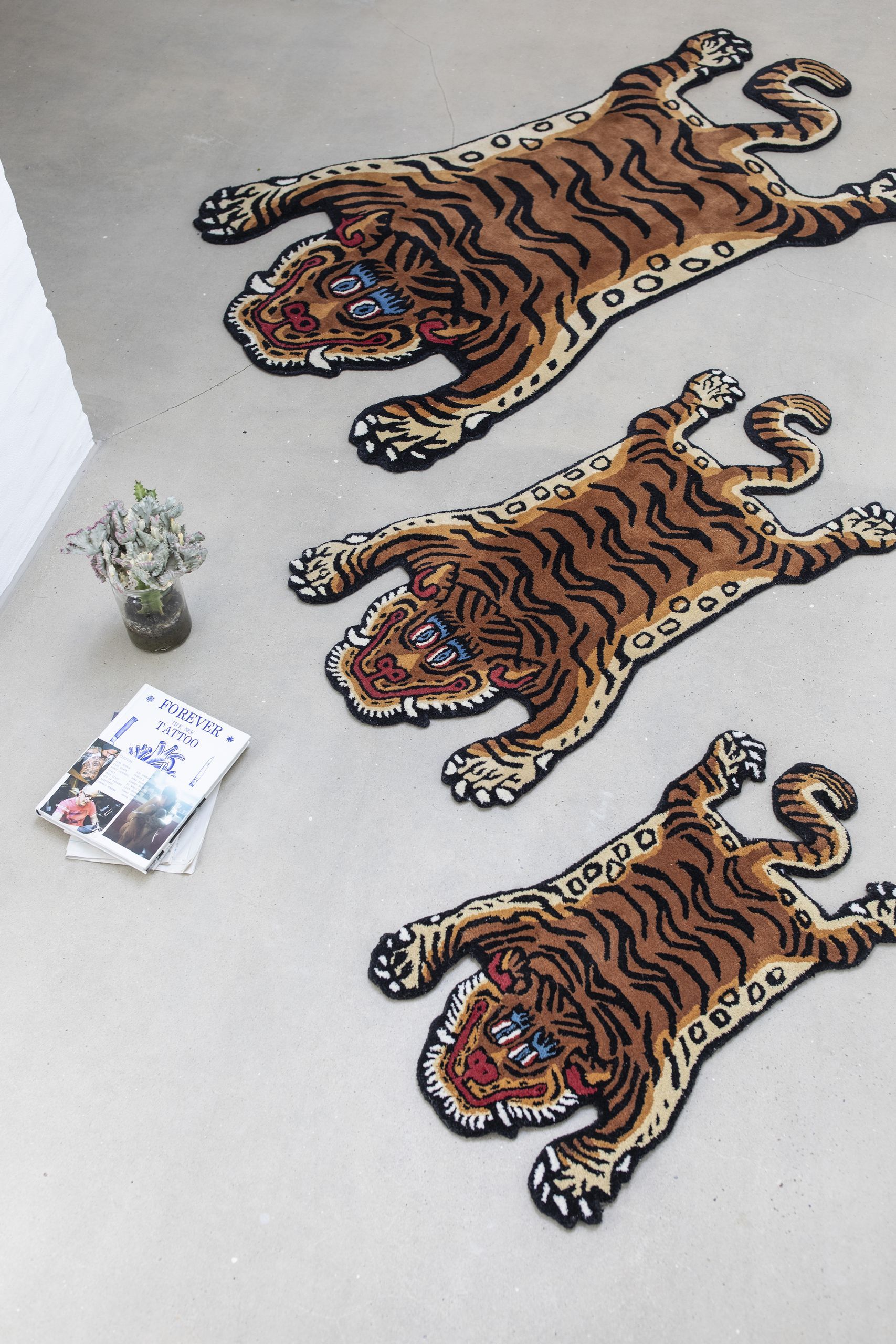Three Bongusta Tiger Rug, Baby rugs are laid on a gray floor near a potted plant and magazines. Arranged diagonally, they add playful jungle vibes with their tiger design and forward-facing heads. Brand: Bongusta.