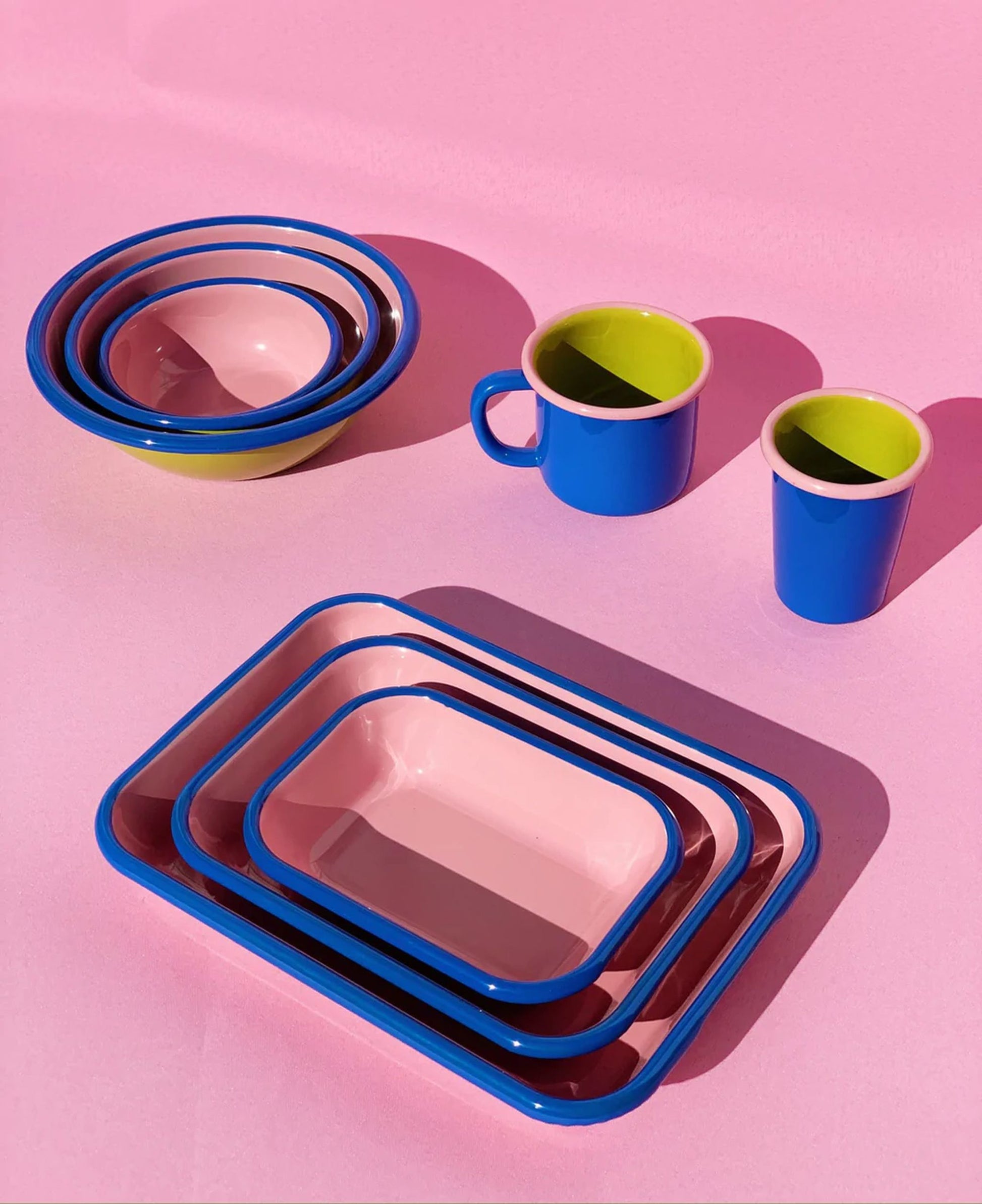 A handmade Bornn Enamelware Colorama Baking Dish, Small, by Bornn Enamelware, features colorful enamel with blue rims and green interiors—seen here on a pink surface with matching bowls and mugs.