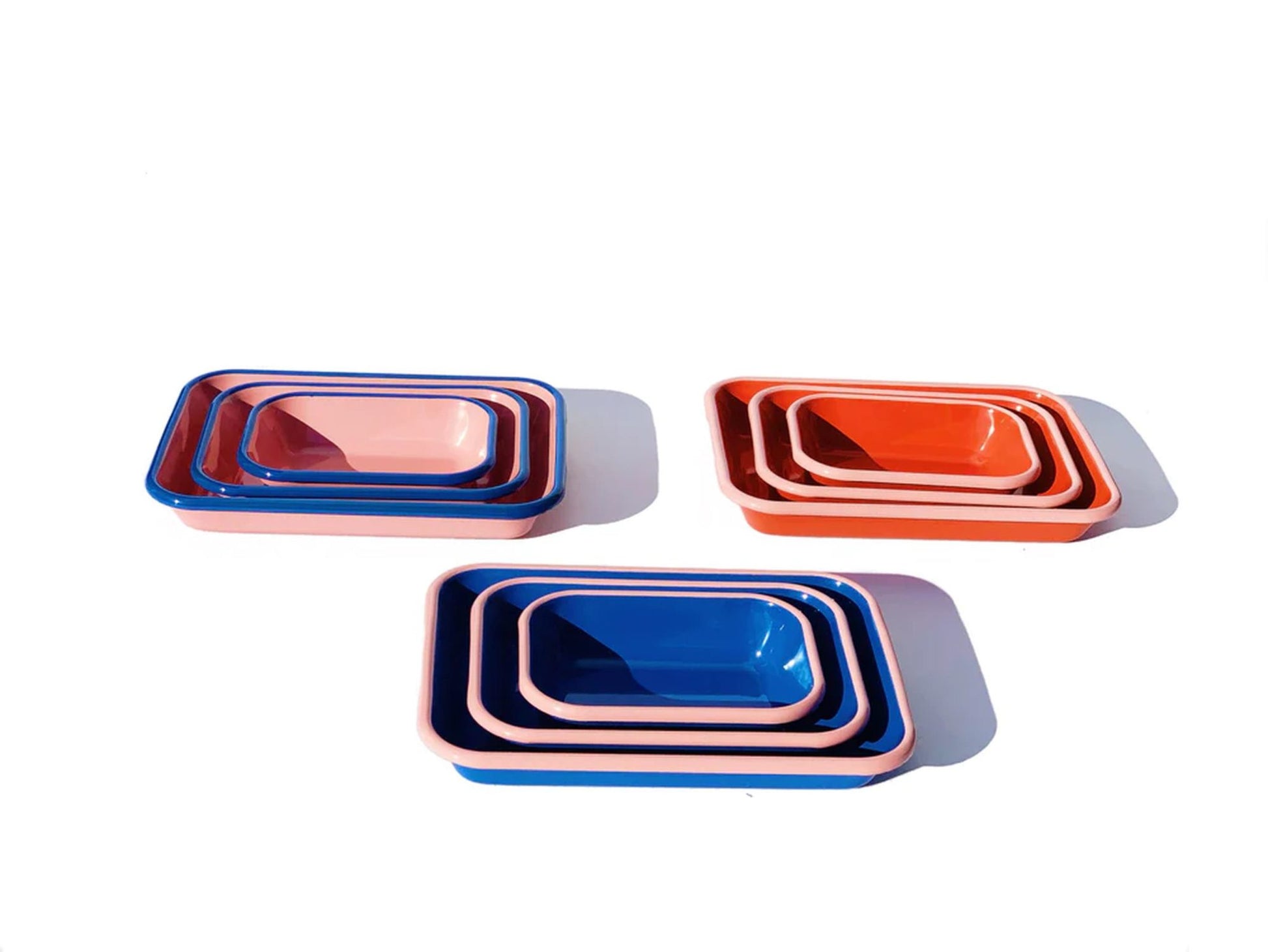 Three Bornn Enamelware Colorama Baking Dish sets in pink, red, and blue are arranged on a white background with soft shadows. Each set by Bornn Enamelware includes four nested trays in decreasing sizes.