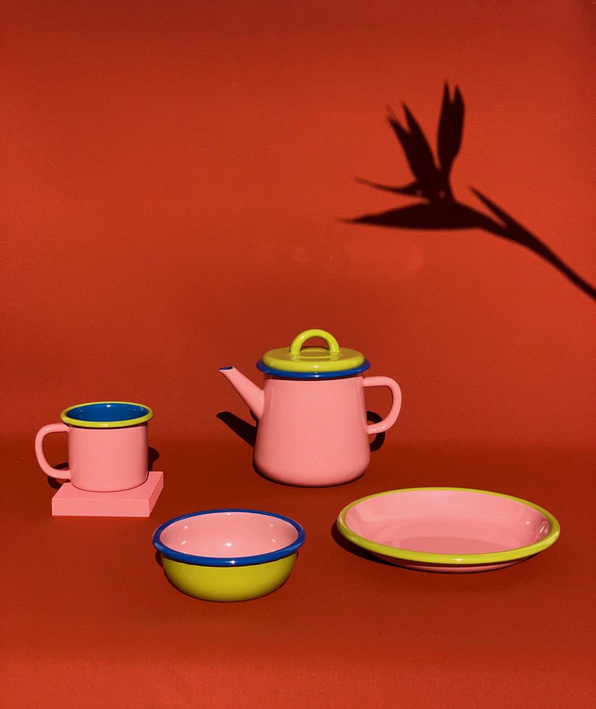 The Bornn Enamelware Colorama Mug by Bornn Enamelware sits on a red surface with a plants shadow in the background, alongside matching pink enamelware teapot, bowl, and plate featuring blue and green rims.