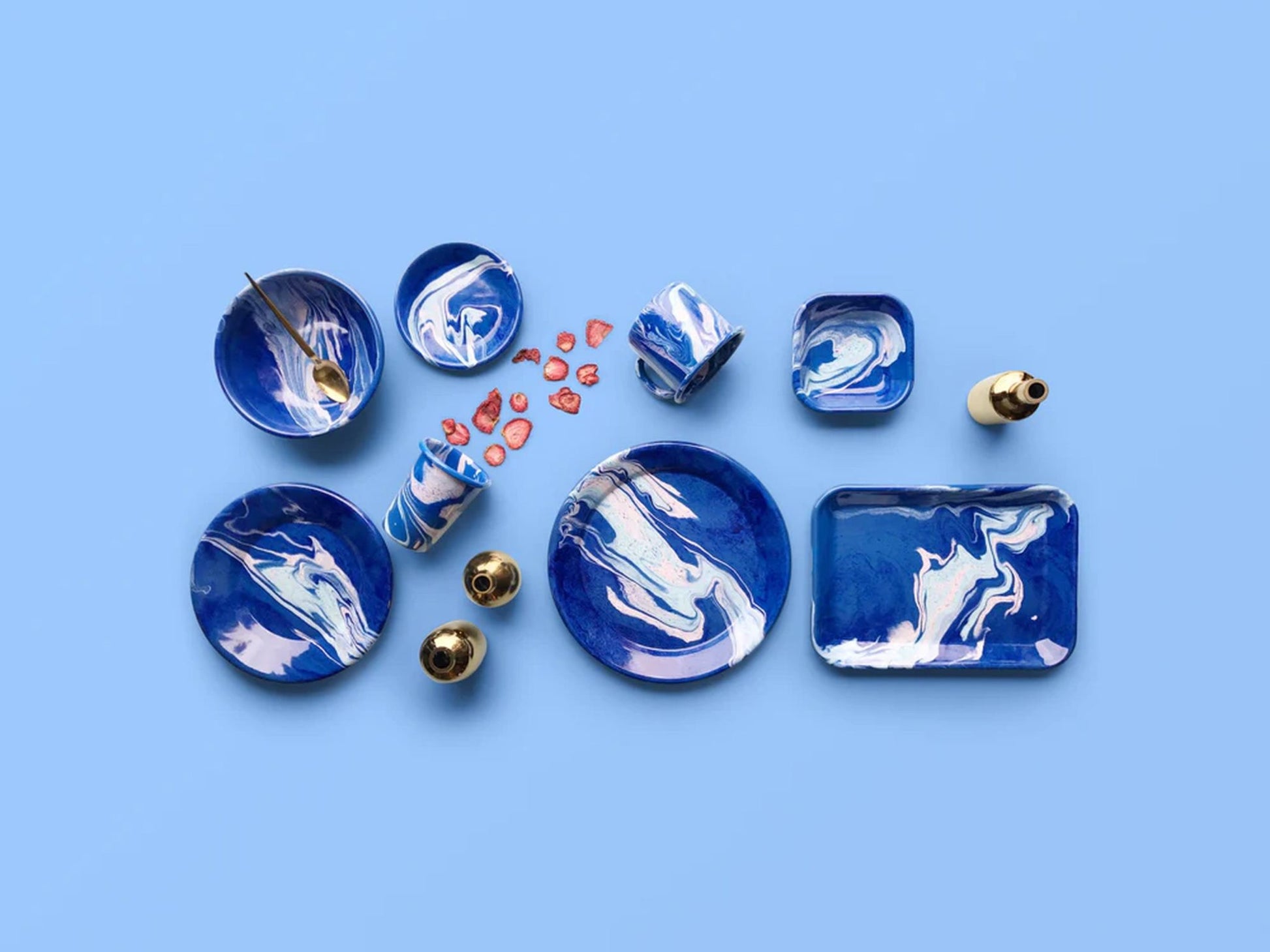 A set of blue and white marbled ceramic dishes from The Stacked Store is arranged with the Bornn Enamelware New Marble Tumbler Cup, gold accents, a bottle, and sliced strawberries on a light blue background.