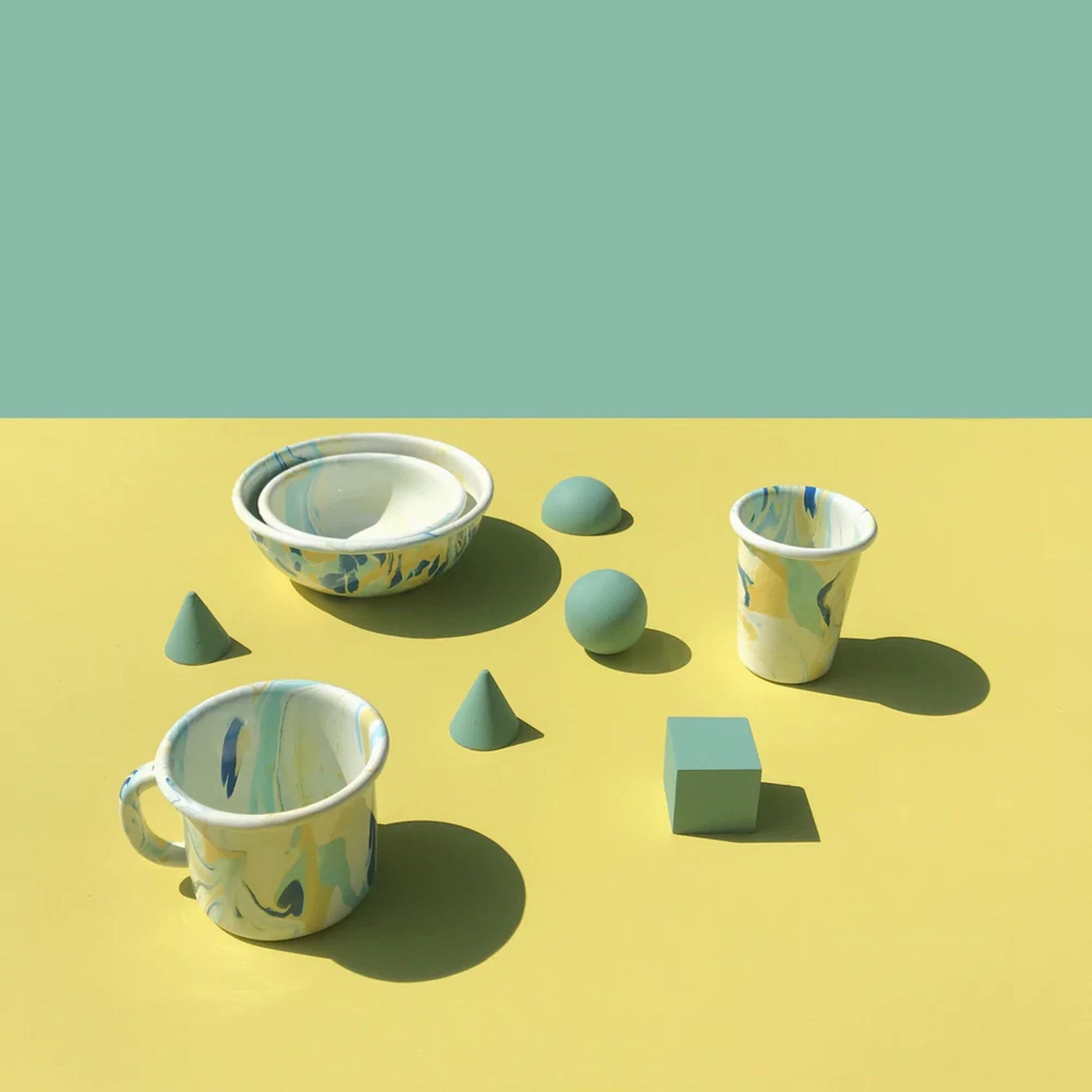 A set of marbled ceramic dishes—bowl, mug, and Bornn Enamelware New Marble Tumbler Cup—by Bornn Enamelware from The Stacked Store, arranged with green geometric shapes on a yellow surface and green background.
