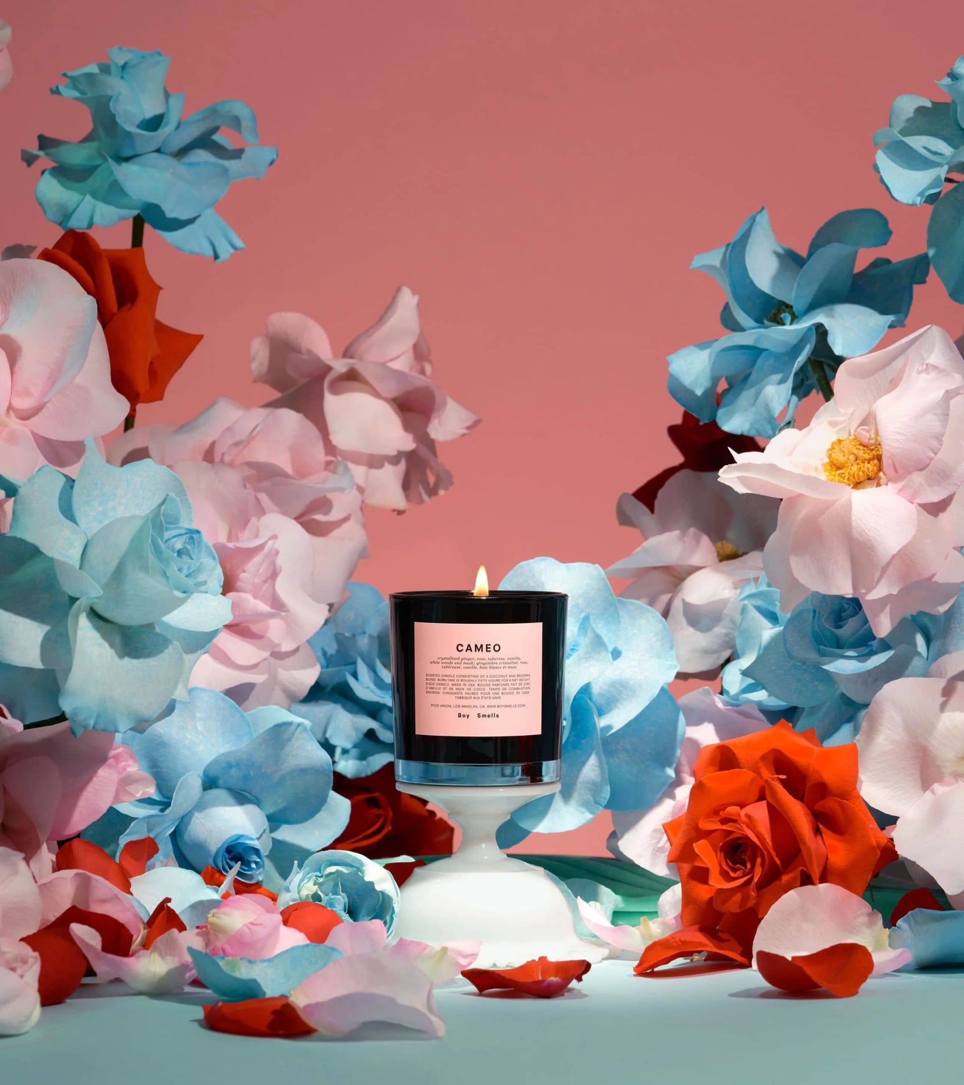 The Boy Smells Cameo Scented Candle 8.5oz, with a pink label, glows at the center, surrounded by bold paper flowers in blue, pink, and red tones against a pink and blue gradient backdrop.