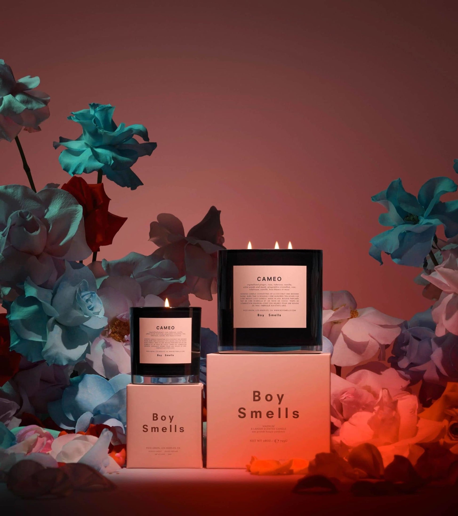 Three Boy Smells Cameo Scented Candle 8.5oz candles glow among vibrant paper flowers under red and blue lights, their floral fragrance filling the air as two rest on boxes and one shines in front, all with their flames lit.