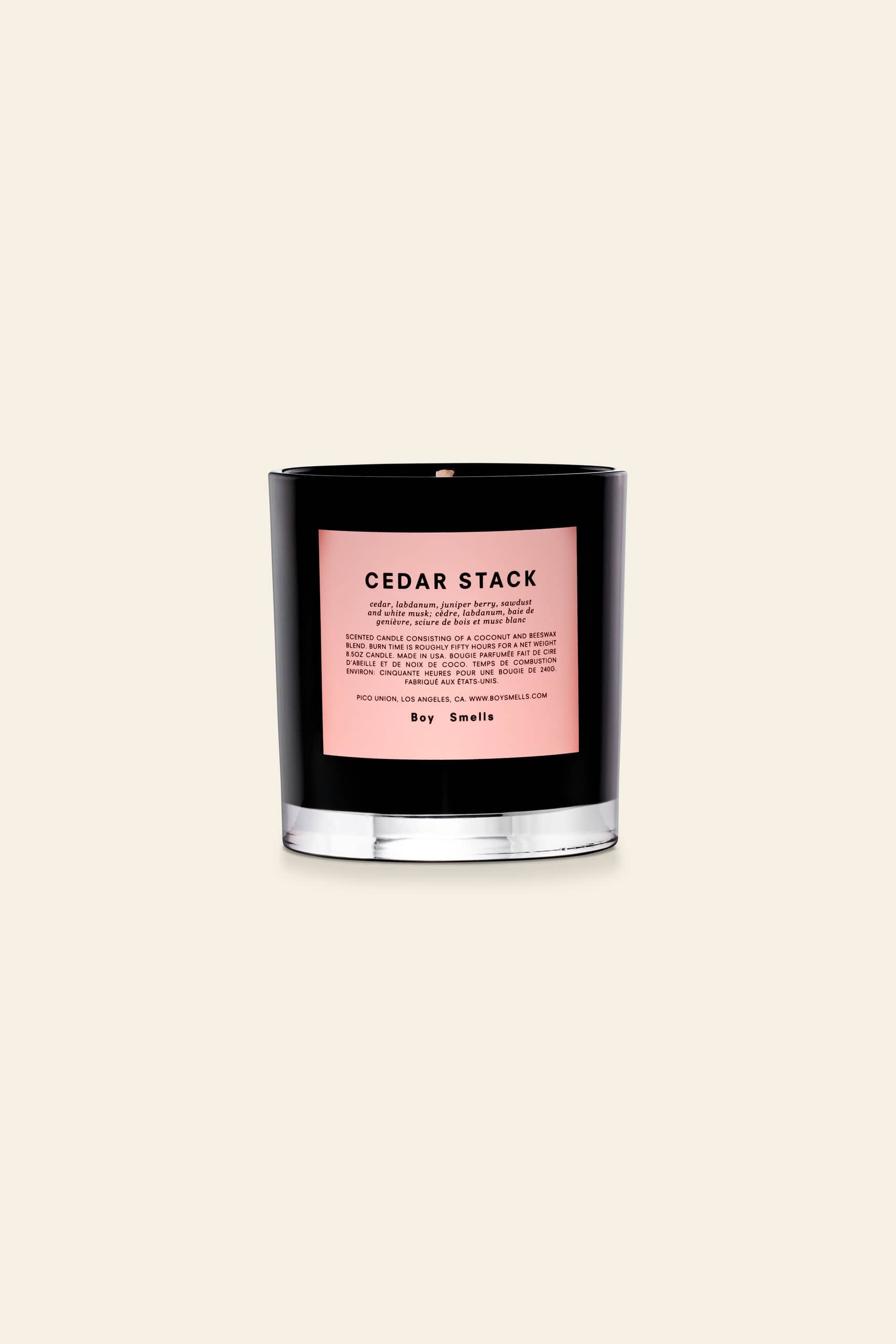 A black glass Boy Smells Cedar Stack Scented Candle 8.5oz jar with a pink label and description sits against a light beige background, highlighting the modern style of this signature candle from Boy Smells.