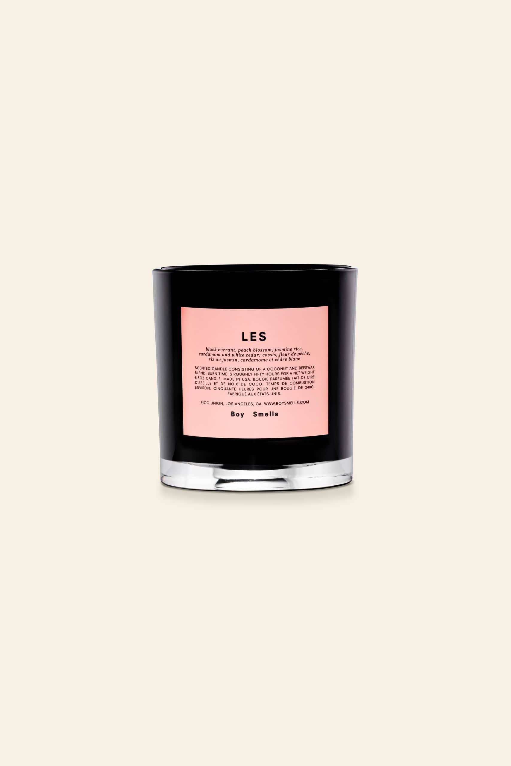 The Boy Smells Les Scented Candle 8.5oz features a black glass jar with a bold pink LES label and product details, plus Boy Smells branding at the bottom. Set against a beige background, it’s an ideal gift for lovers of unique fragrances.