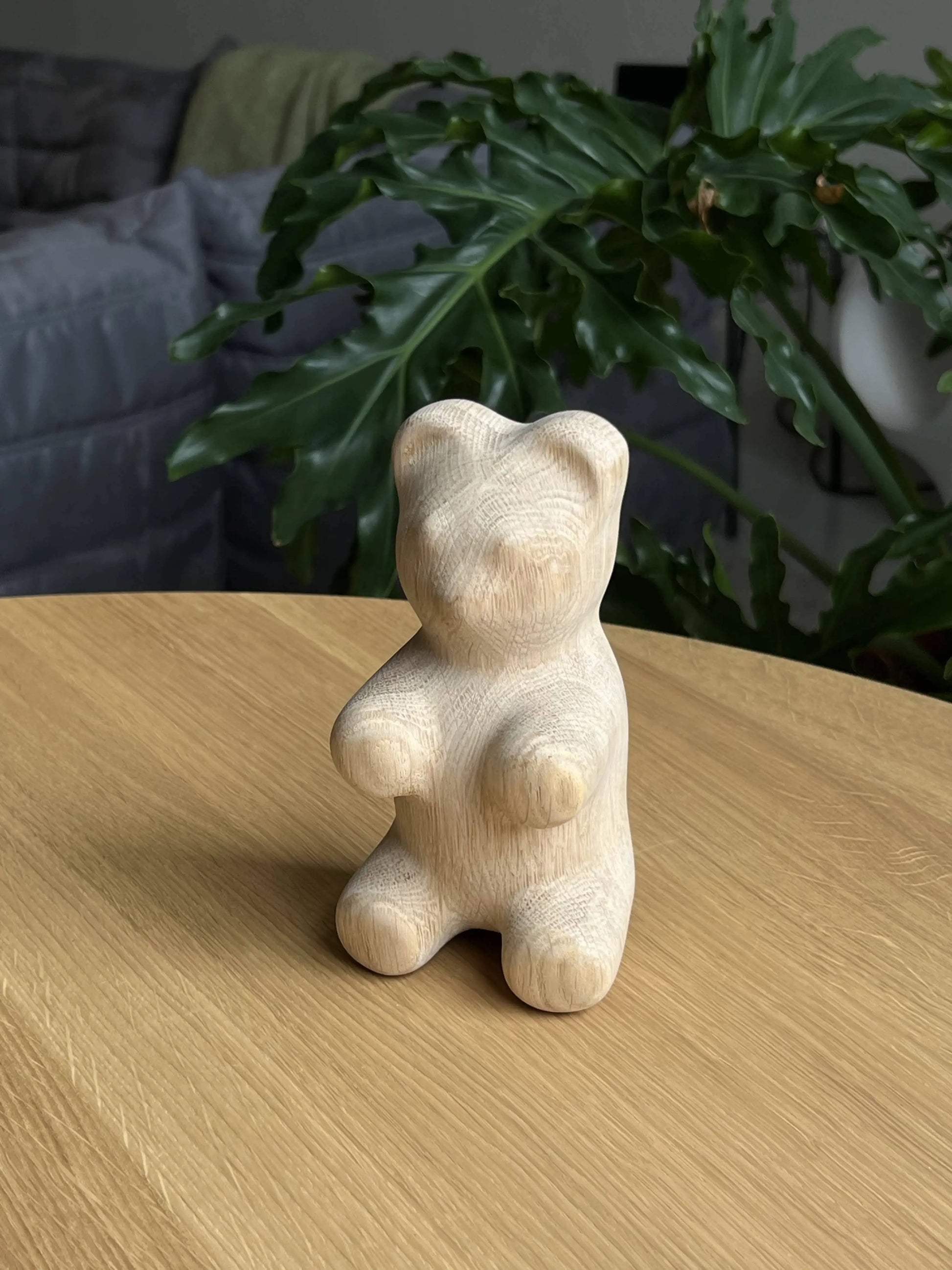 The Boyhood Gummy Bear Small by Boyhood, a smooth wooden bear figurine, sits on a light wooden table amid green leafy plants and a gray couch softly blurred in the background.