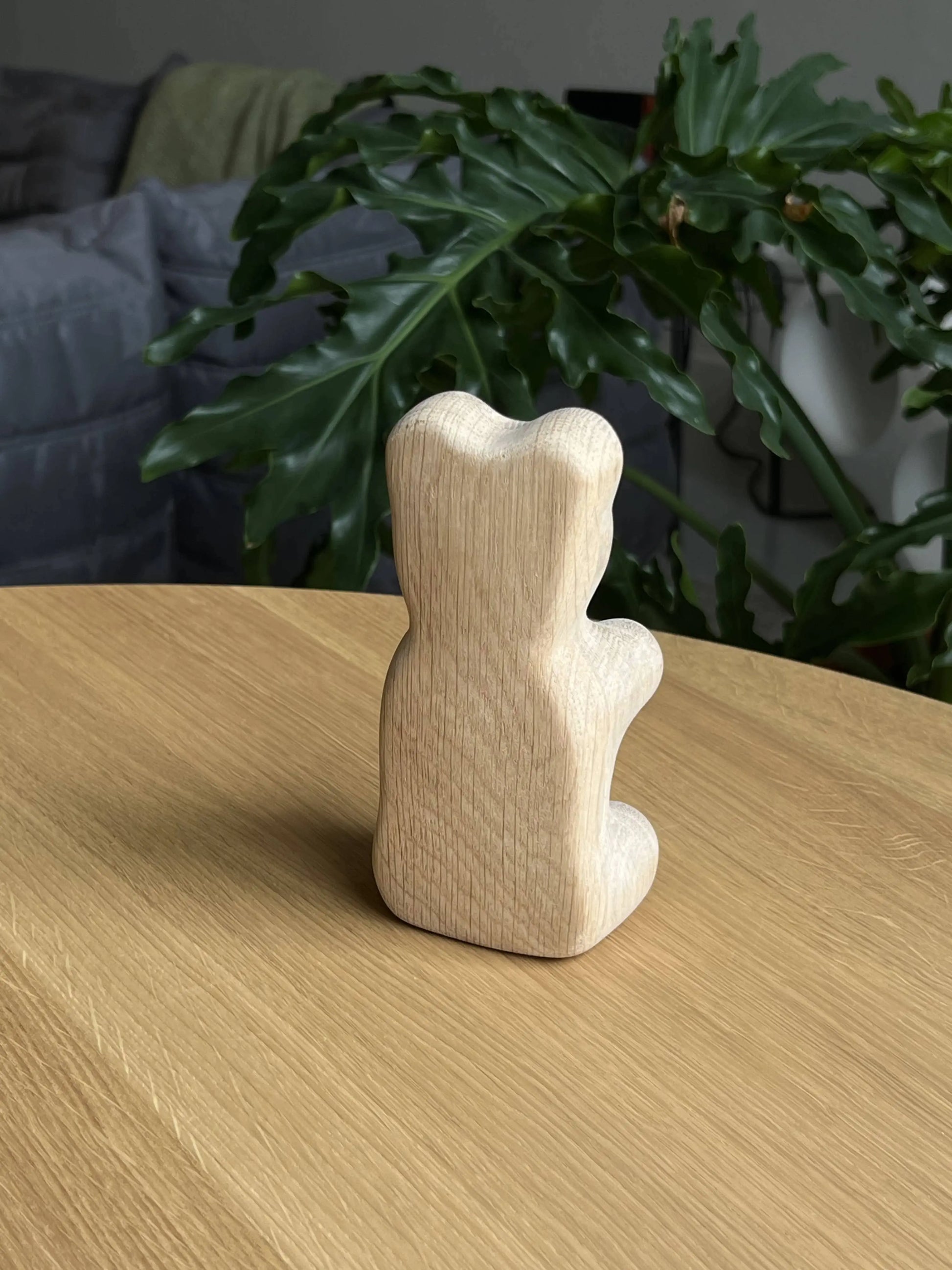 The Boyhood Gummy Bear Small by Boyhood, an unfinished wooden figurine inspired by the classic candy, sits on a light wood table facing away. A green leafy plant and gray sofa in the background add subtle charm to the scene.