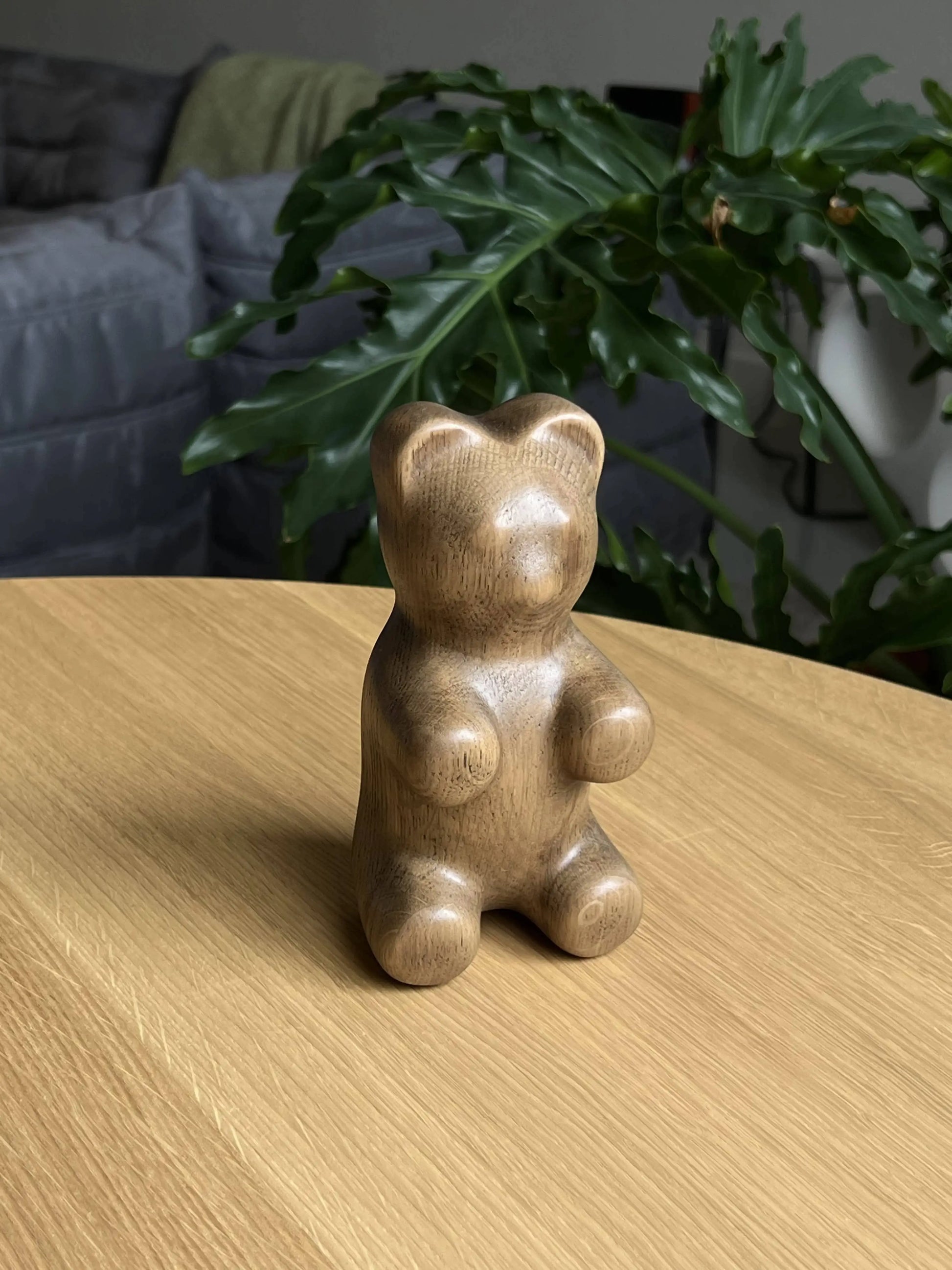 The Boyhood Gummy Bear Small by Boyhood, a smooth wooden figurine, rests on a light wood table with a green leafy plant and part of a gray couch in the background.