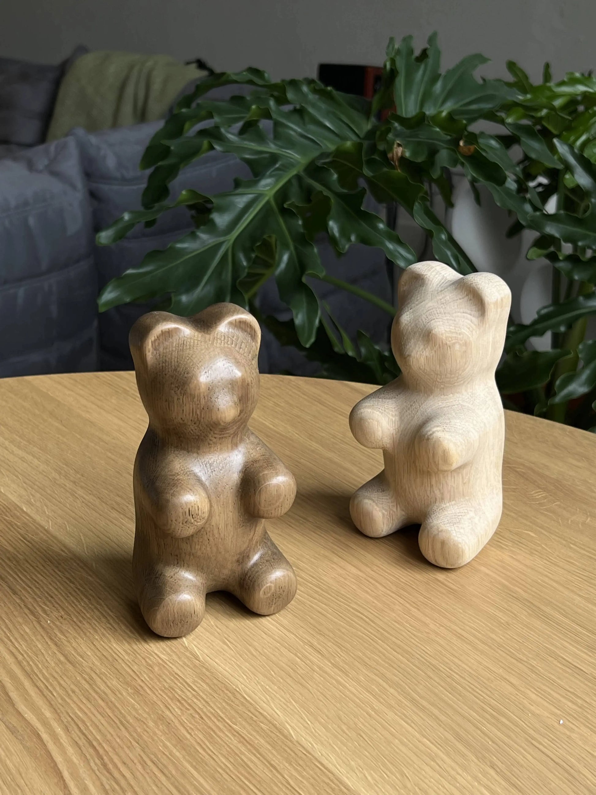 Two Boyhood Gummy Bear Small figurines by Boyhood, one dark brown and one light beige, are displayed on a wooden table with a green leafy plant and part of a gray sofa in the background, adding charm to any space.