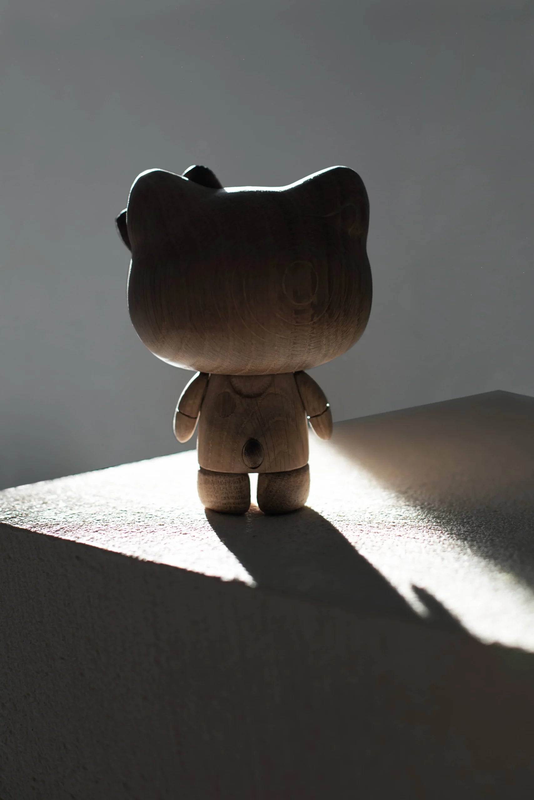 The Boyhood Hello Kitty Small figurine by Boyhood, a charming wooden collectible, stands on a sunlit surface casting a long shadow. Viewed from behind, dramatic light accentuates its smooth wooden texture.