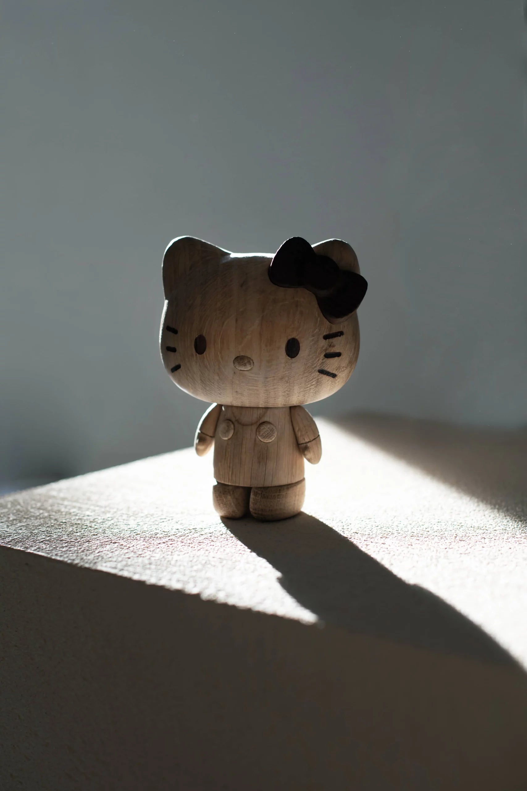The Boyhood Hello Kitty Small by Boyhood features a wooden figurine with a black bow, standing on a sunlit surface and casting a long shadow—a charming collectible for any Hello Kitty or wooden figure enthusiast.