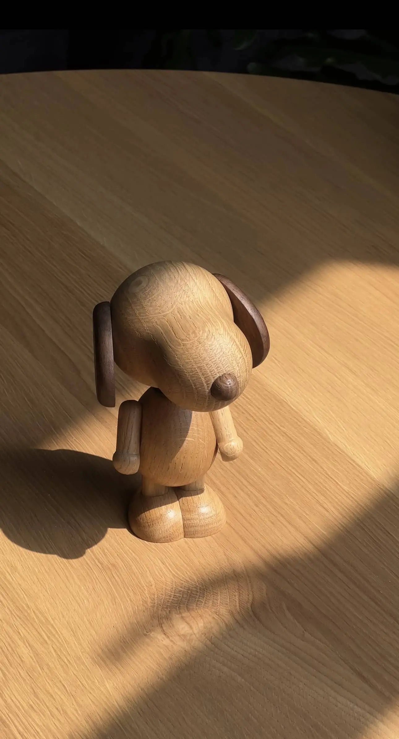 The Boyhood Snoopy Small by Boyhood, a wooden dog figurine with dark brown ears and nose, stands on a light wood surface, partially lit by sunlight—evoking Snoopy enjoying the outdoors.