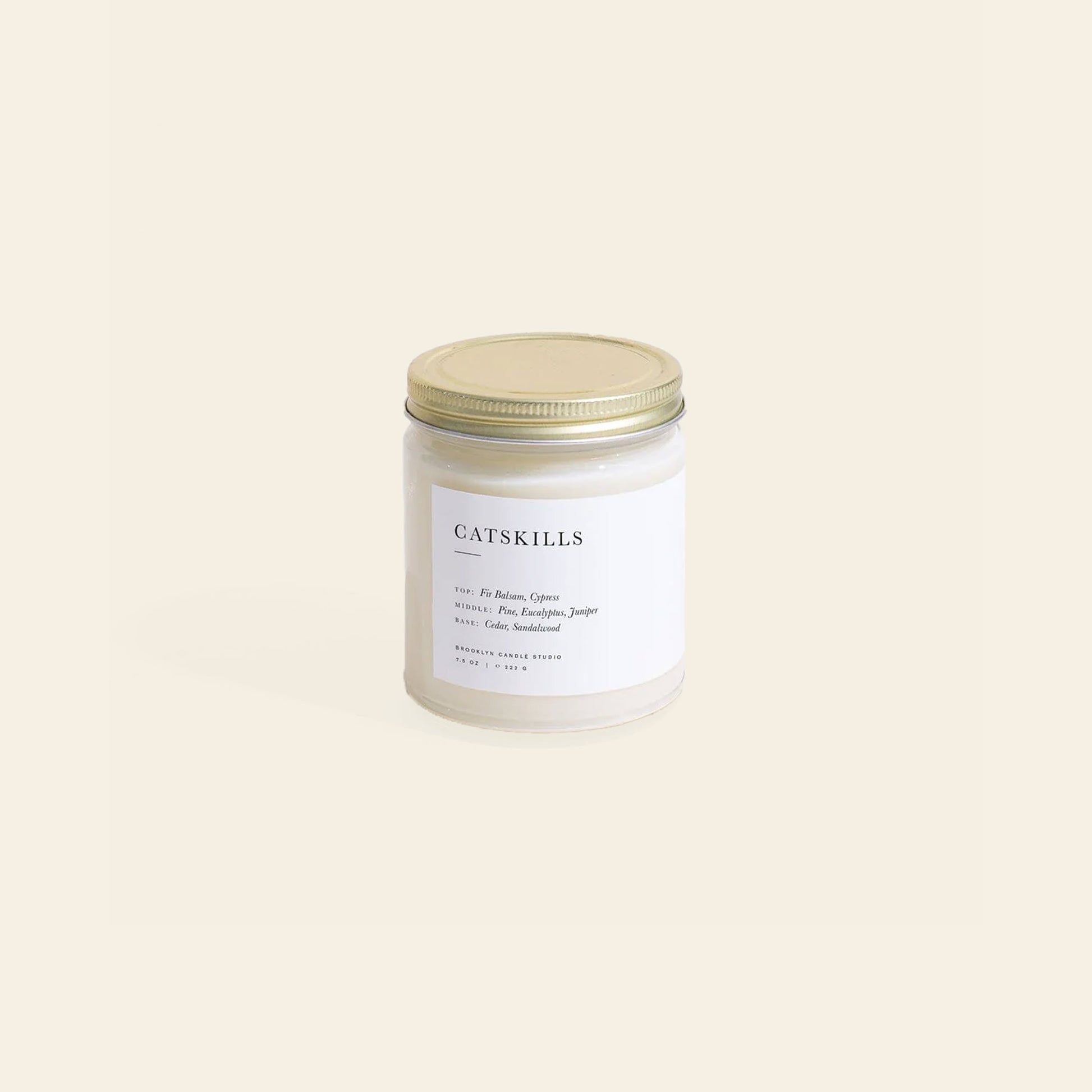 The Brooklyn Candle Studio Catskills Jar Candle features a glass jar with a gold lid and a white label displaying CATSKILLS, woodsy fragrance notes, and brand details. Set against a light beige background, it evokes a calm ambiance.