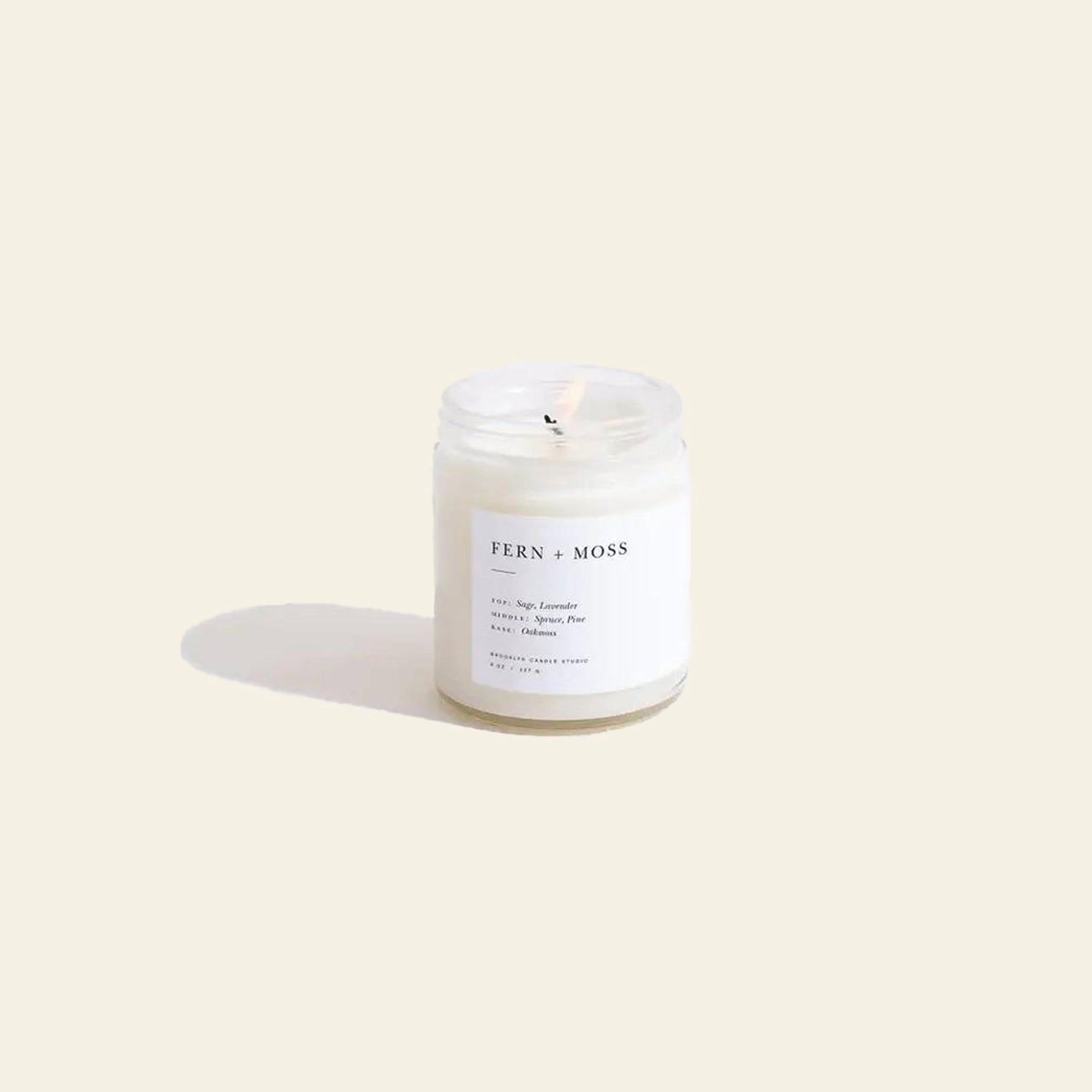 The Brooklyn Candle Studio Fern + Moss Jar Candle rests on a light beige surface. Housed in clear glass with a minimalist label, this single-wick candle offers a nature-inspired scent crafted from essential oils.