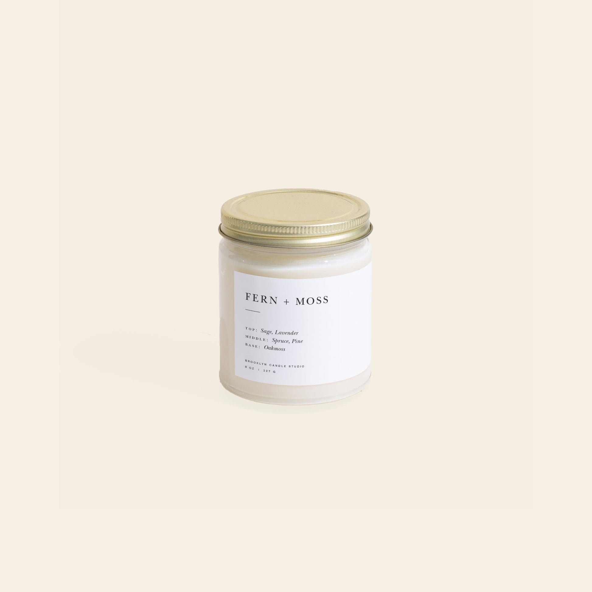 The Brooklyn Candle Studio Fern + Moss Jar Candle features a gold lid and rests on a light beige background. Its white label in minimal black text lists sage, lavender, and orange—nature-inspired notes crafted with essential oils.