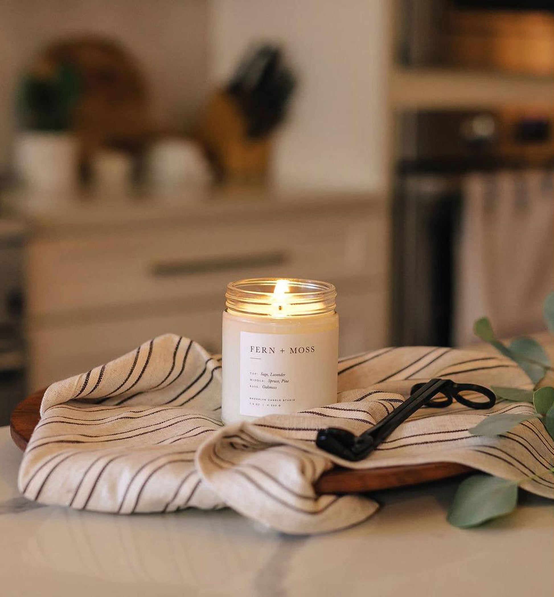 The Brooklyn Candle Studio Fern + Moss Jar Candle, known for its nature-inspired scent, is displayed on a striped cloth with a wick trimmer on a wooden tray atop a kitchen counter, with blurred kitchen items in the background.